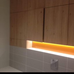 Bartłomiej Maczuga - Nowoczesna łazienka z drewnianymi szafkami wiszącymi i podświetleniem LED nad szarą, perforowaną ścianą z płytek oraz chromowaną armaturą.