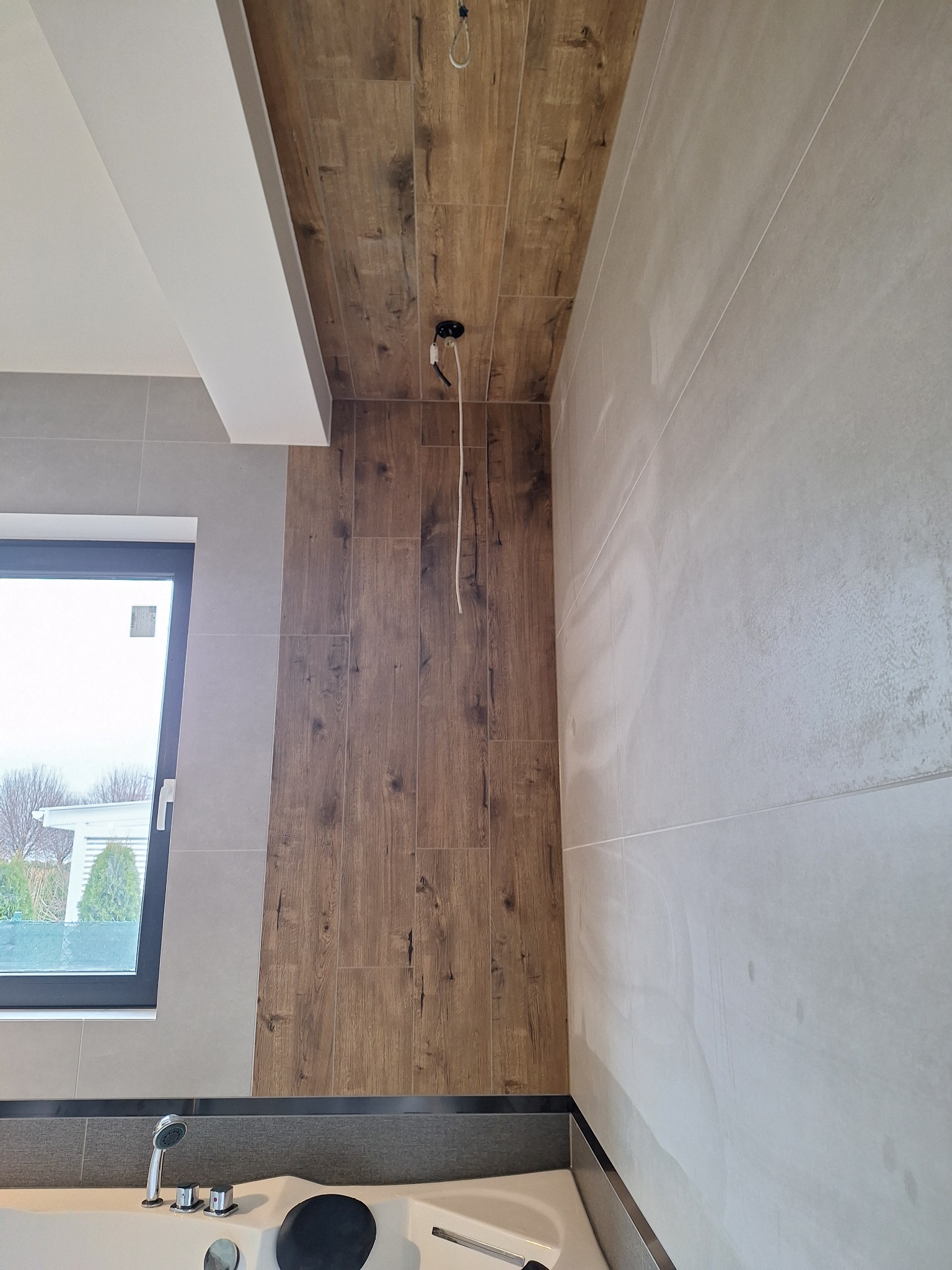 Wnętrze łazienki w trakcie wykańczania: połączenie płytek imitujących drewno na ścianie i suficie z jasnoszarymi płytkami, widoczna wanna z hydromasażem i okno.