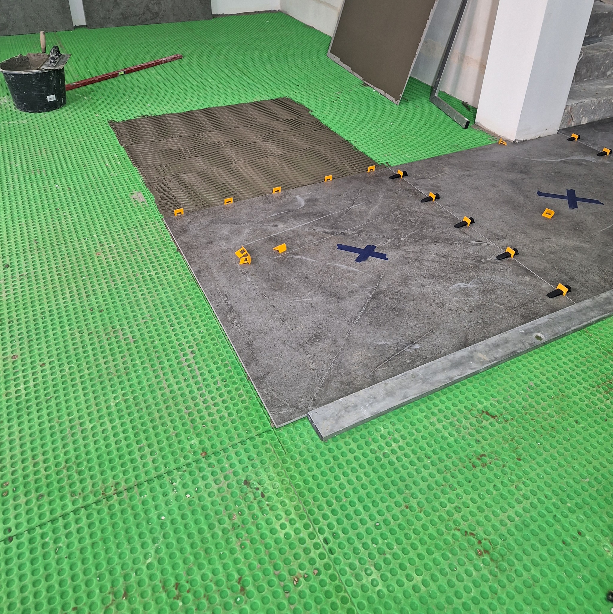 Szare płytki podłogowe układane na zielonej membranie izolacyjnej, z użyciem poziomicy i klipsów poziomujących.