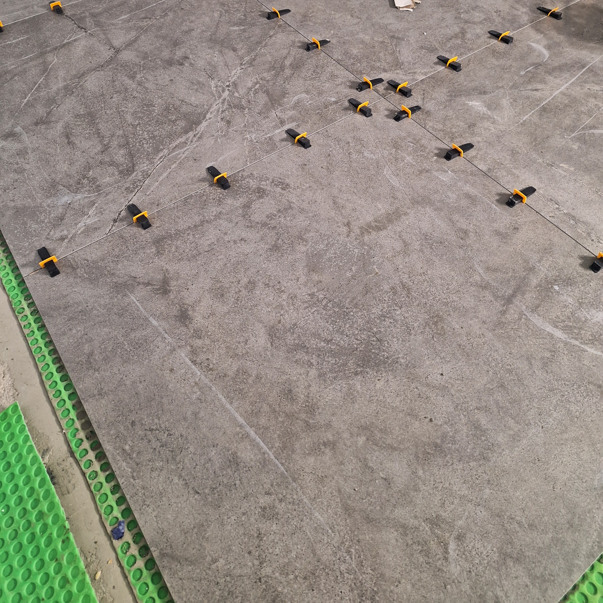 Szara płytka podłogowa imitująca kamień, z widocznymi systemami poziomowania płytek (klipsy i kliny) oraz zieloną matą drenażową przy krawędzi.