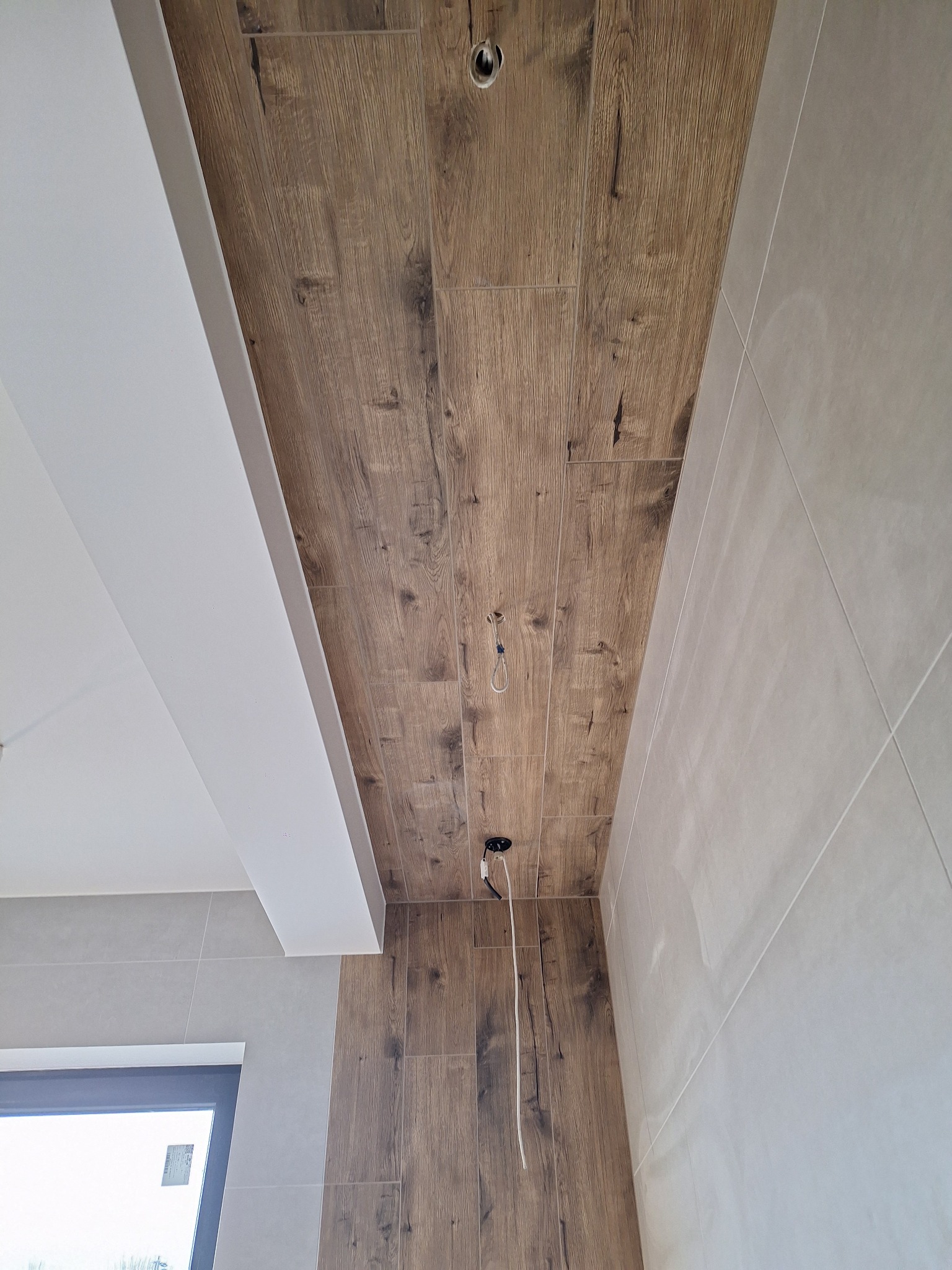 Ściana i sufit wyłożone płytkami imitującymi drewno, z widocznymi otworami na oświetlenie i wystającymi kablami, oraz fragment ściany z szarymi płytkami i oknem w tle.