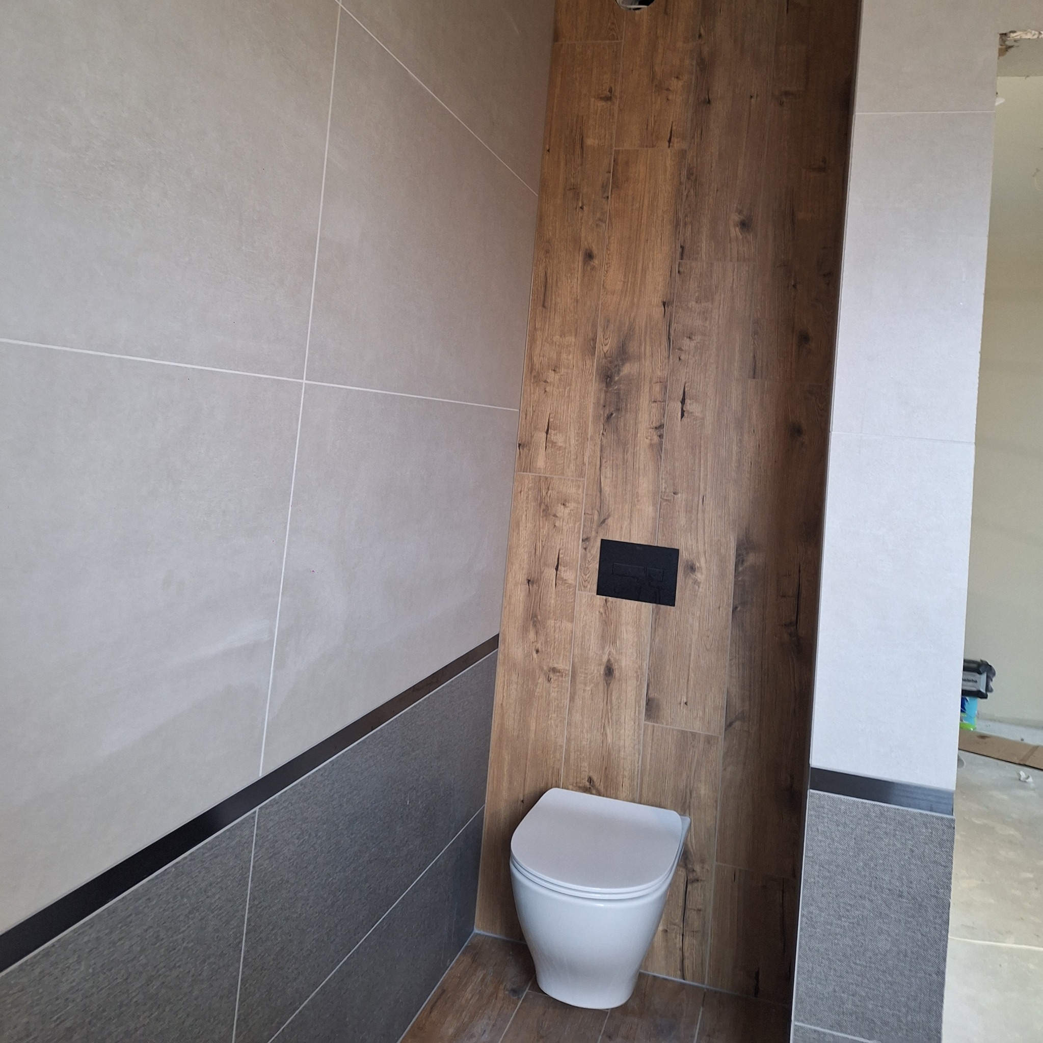 Fragment wykończonej łazienki z podwieszaną toaletą, drewnopodobną okładziną ściany i jasnymi, wielkoformatowymi płytkami na ścianach, z widocznym czarnym panelem sterującym spłuczką.