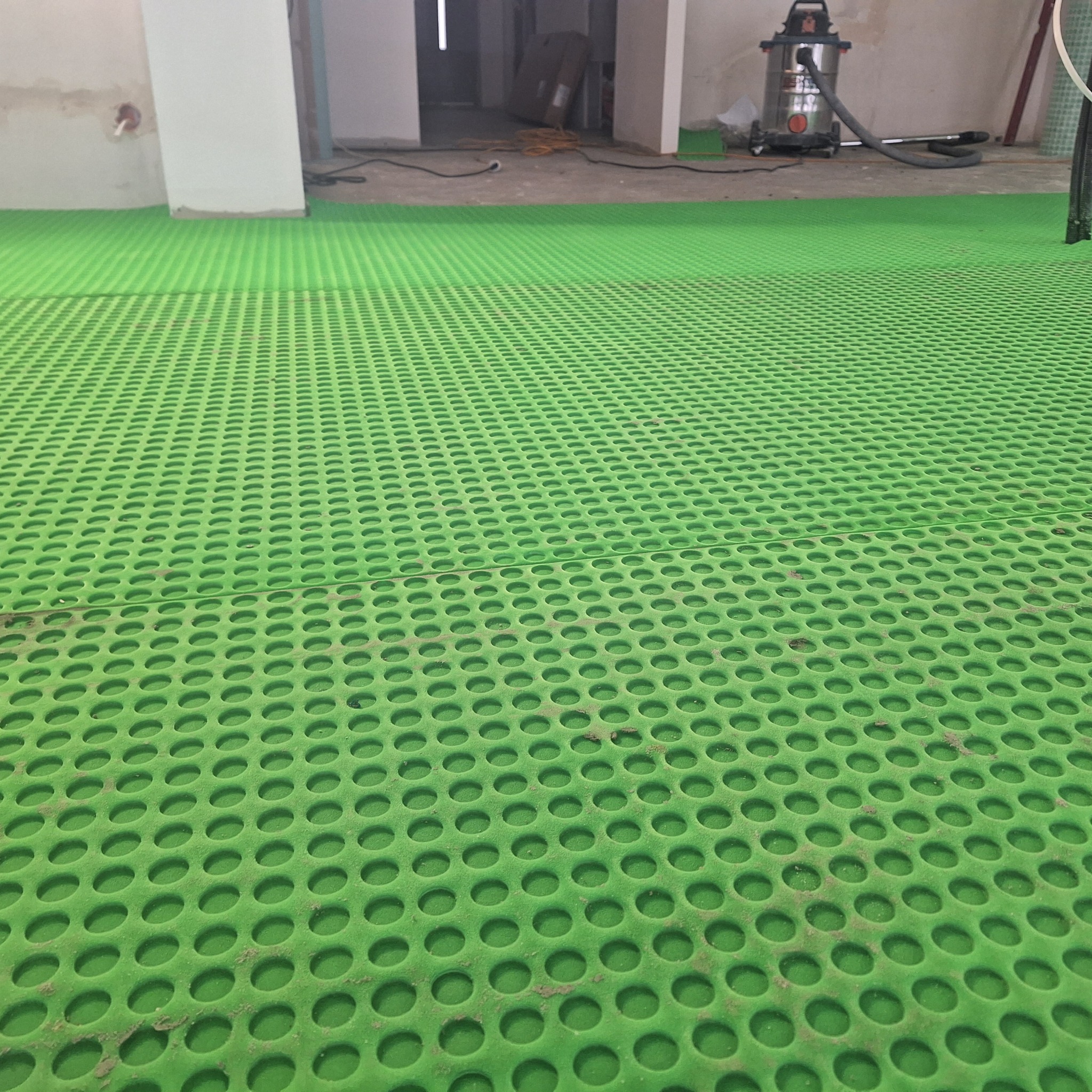 Zielona, perforowana mata podłogowa w trakcie montażu w pomieszczeniu w stanie surowym, widoczny odkurzacz przemysłowy i przewody.