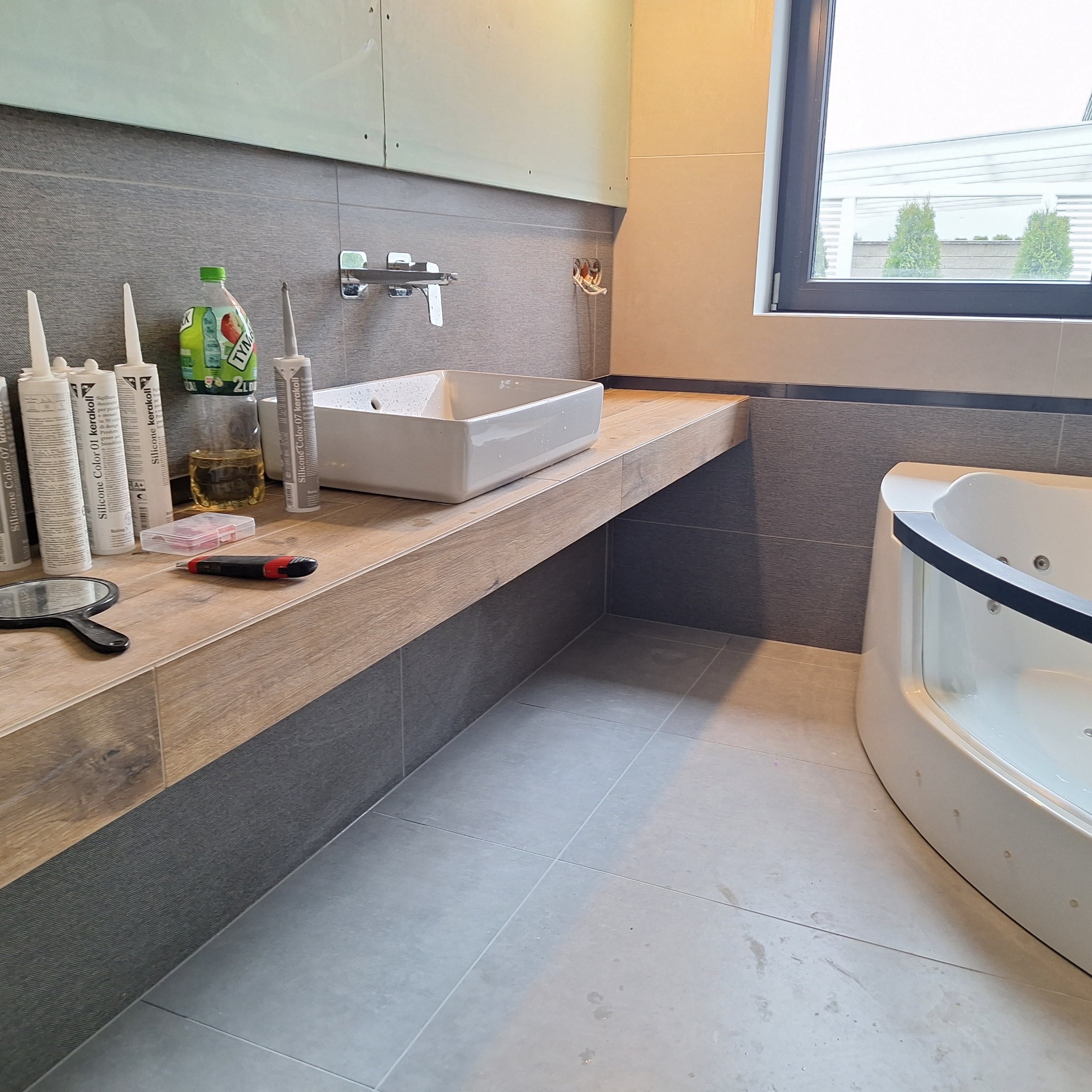 Łazienka w trakcie wykańczania: umywalka nablatowa, silikony, narzędzia i płytki imitujące drewno na blacie oraz szare płytki na ścianach i podłodze, widoczna wanna z hydromasażem i okno z widokiem...
