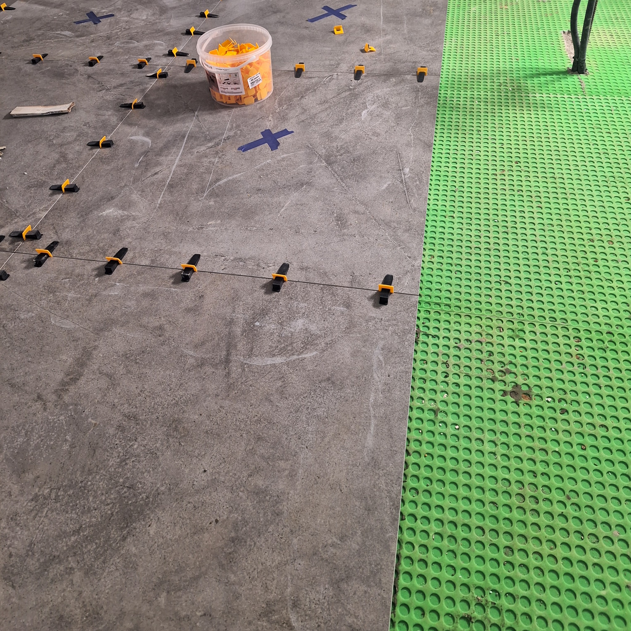 Szare płytki podłogowe w trakcie układania, z użyciem systemu poziomowania płytek (klipsy i kliny), widoczny także pojemnik z klinami i zielona mata ochronna na podłodze.