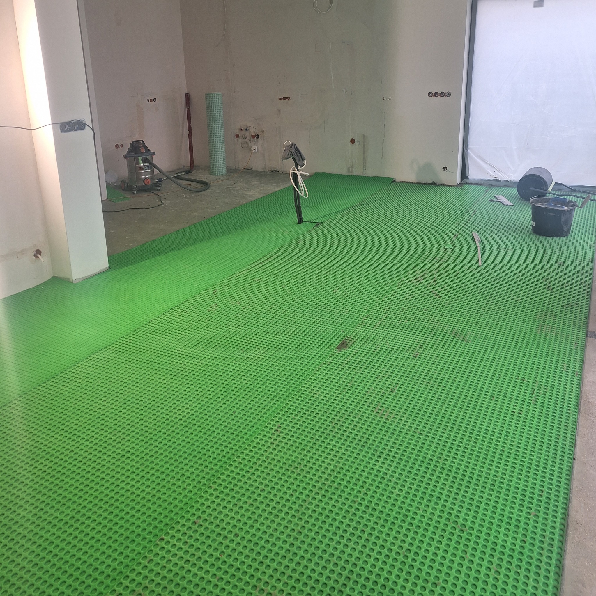 Przygotowanie podłoża pod wylewkę: zielona mata drenażowa z otworami ułożona na surowej podłodze w pomieszczeniu w trakcie remontu, widoczny odkurzacz przemysłowy, wiadro i narzędzia.