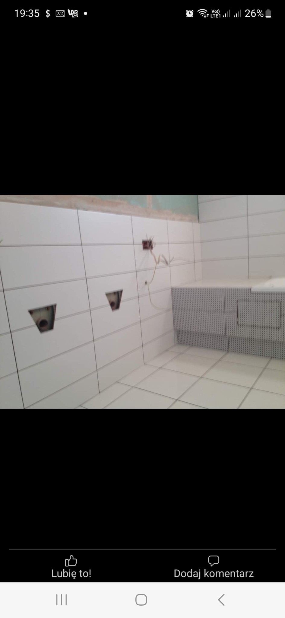 Białe płytki ścienne i podłogowe w trakcie montażu w łazience, widoczne otwory na armaturę i wystające przewody elektryczne, częściowo wykończona zabudowa w rogu pomieszczenia.