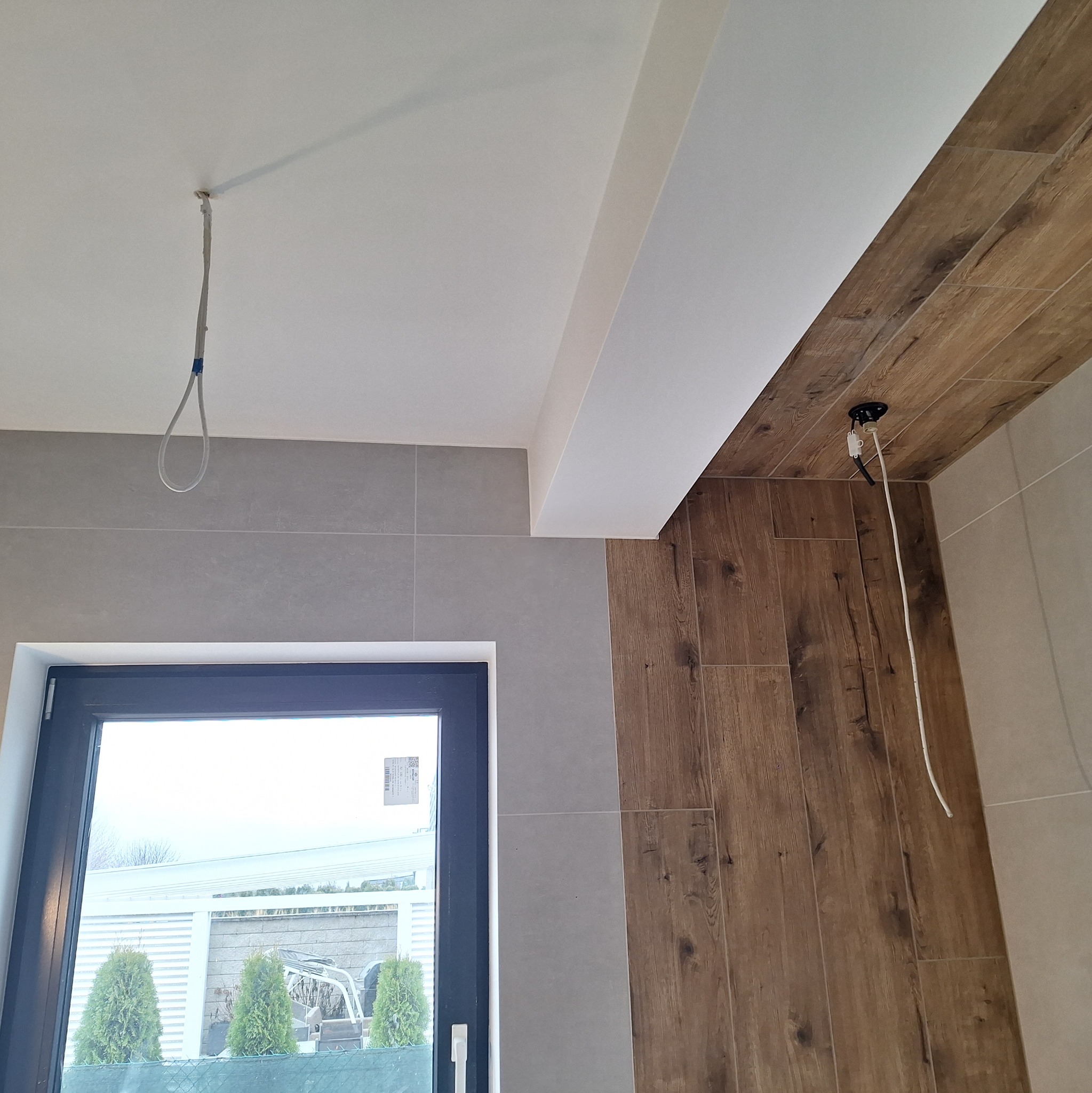 Wnętrze łazienki w trakcie prac wykończeniowych: szare płytki ścienne, drewnopodobne płytki na suficie, wystający kabel elektryczny i okno z widokiem na zewnątrz.