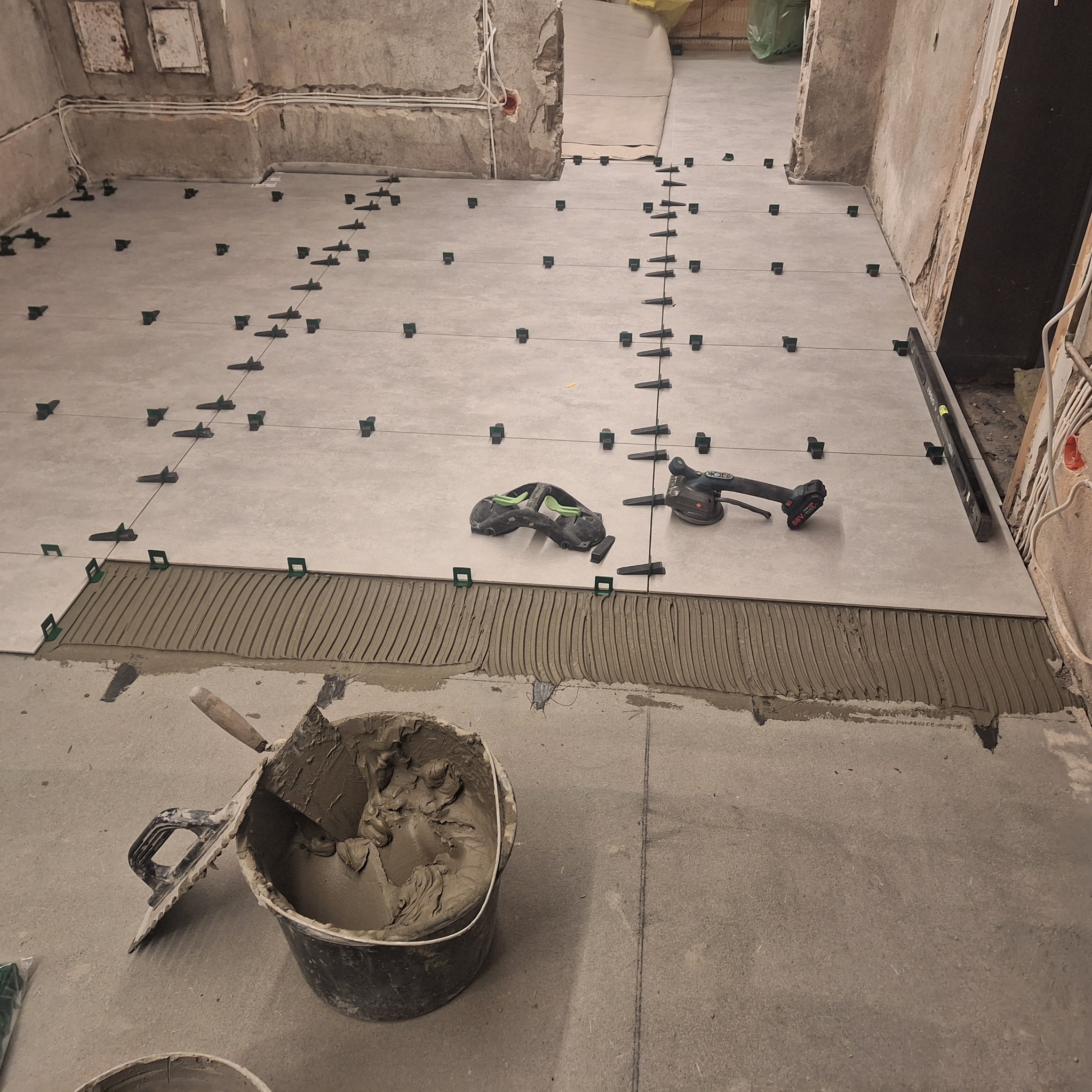 Układanie płytek podłogowych: widok na przygotowany fragment podłogi z nałożonym klejem, poziomica, przyssawka do płytek i wiadro z zaprawą w pomieszczeniu w stanie surowym.