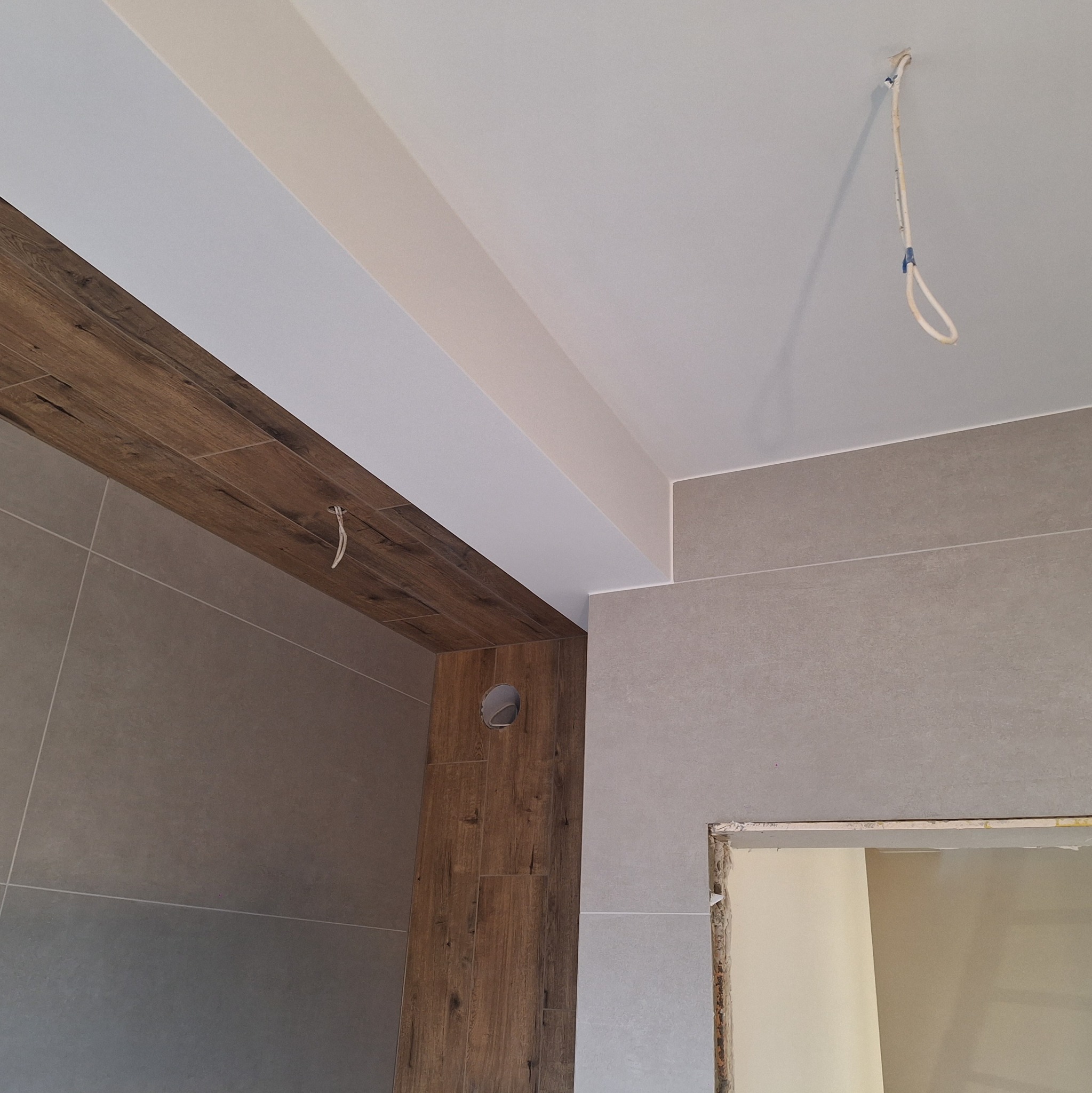 Wnętrze łazienki w trakcie prac wykończeniowych, widoczne szare płytki na ścianach, drewnopodobne płytki na ścianie z otworem wentylacyjnym, biały sufit z wystającymi przewodami elektrycznymi...