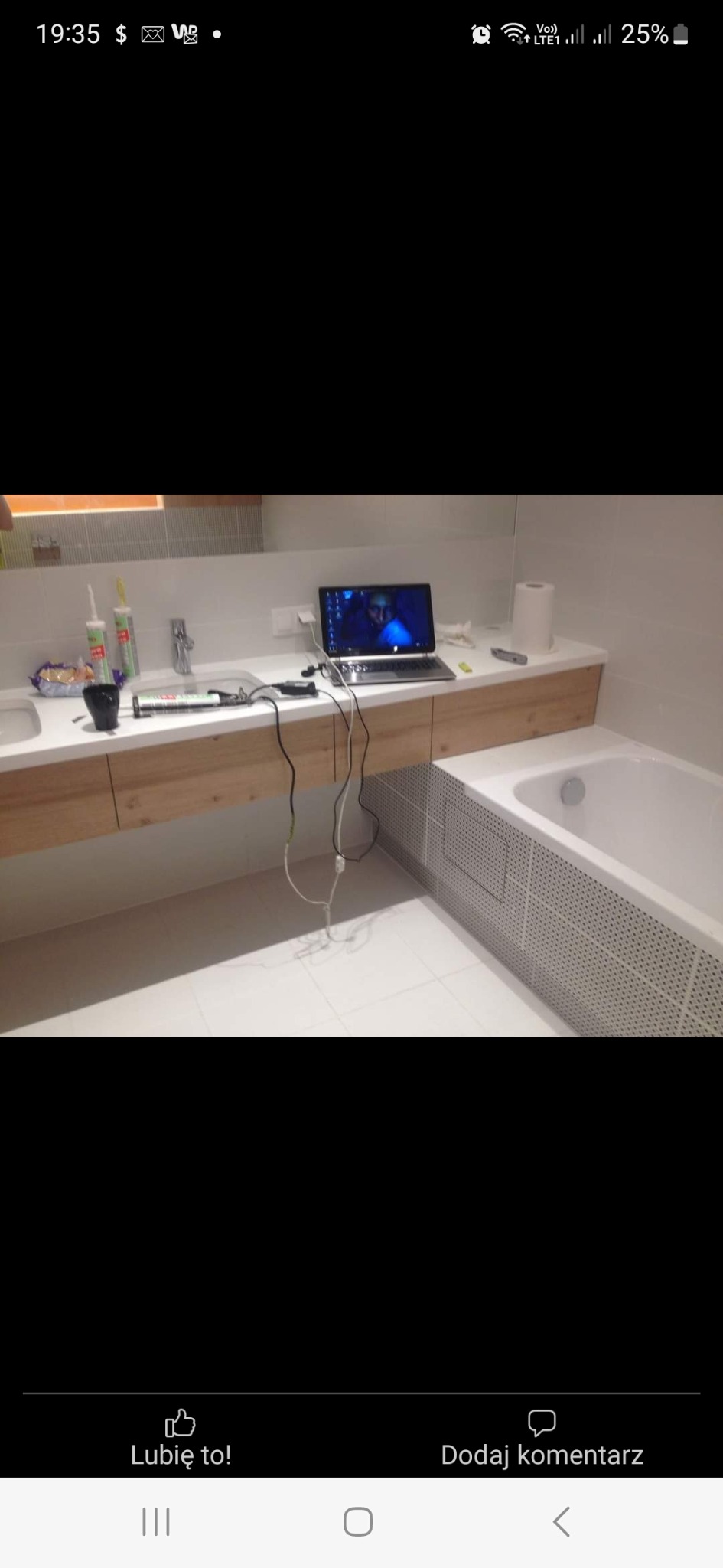 Nowoczesna łazienka z białymi płytkami, drewnianą szafką pod umywalką i wanną obudowaną płytkami z perforacją, na blacie laptop z włączonym ekranem, tuby silikonu i akcesoria.