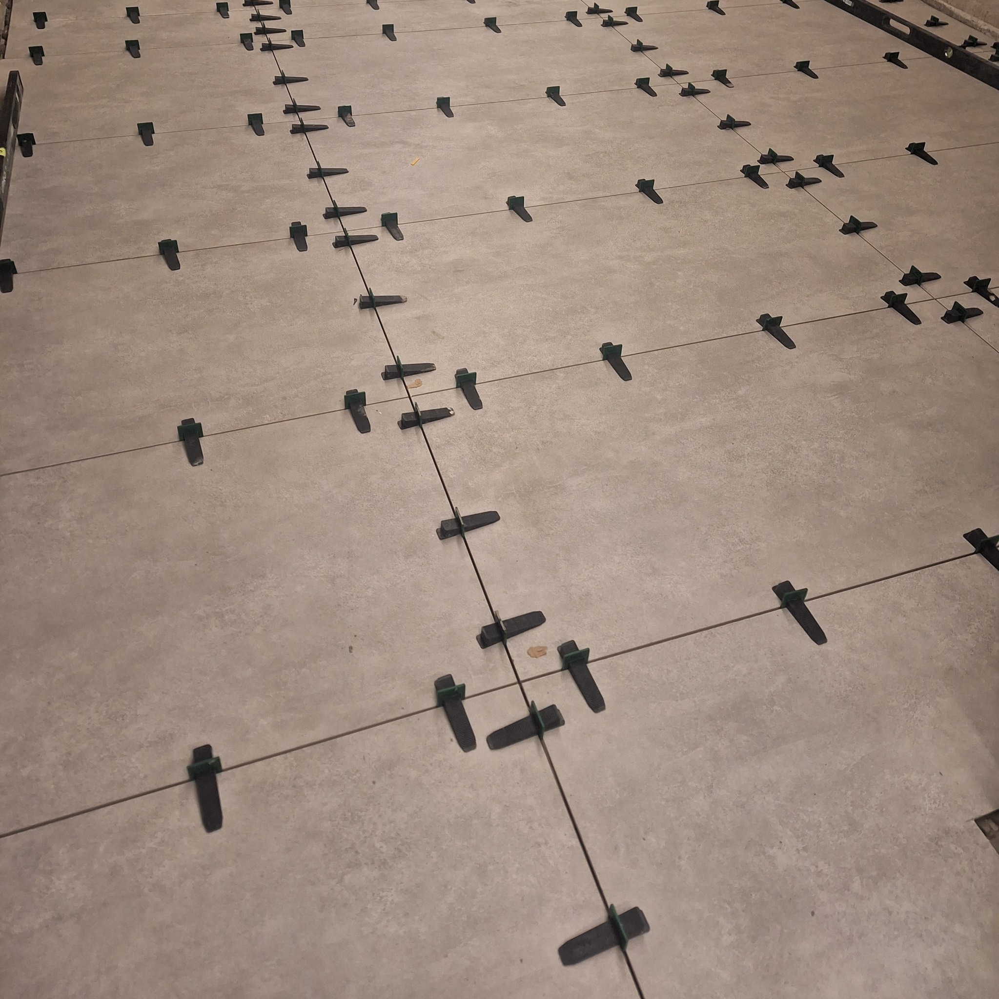 Szare płytki podłogowe w trakcie układania, z użyciem systemu poziomowania płytek, widoczne krzyżyki dystansowe i kliny.