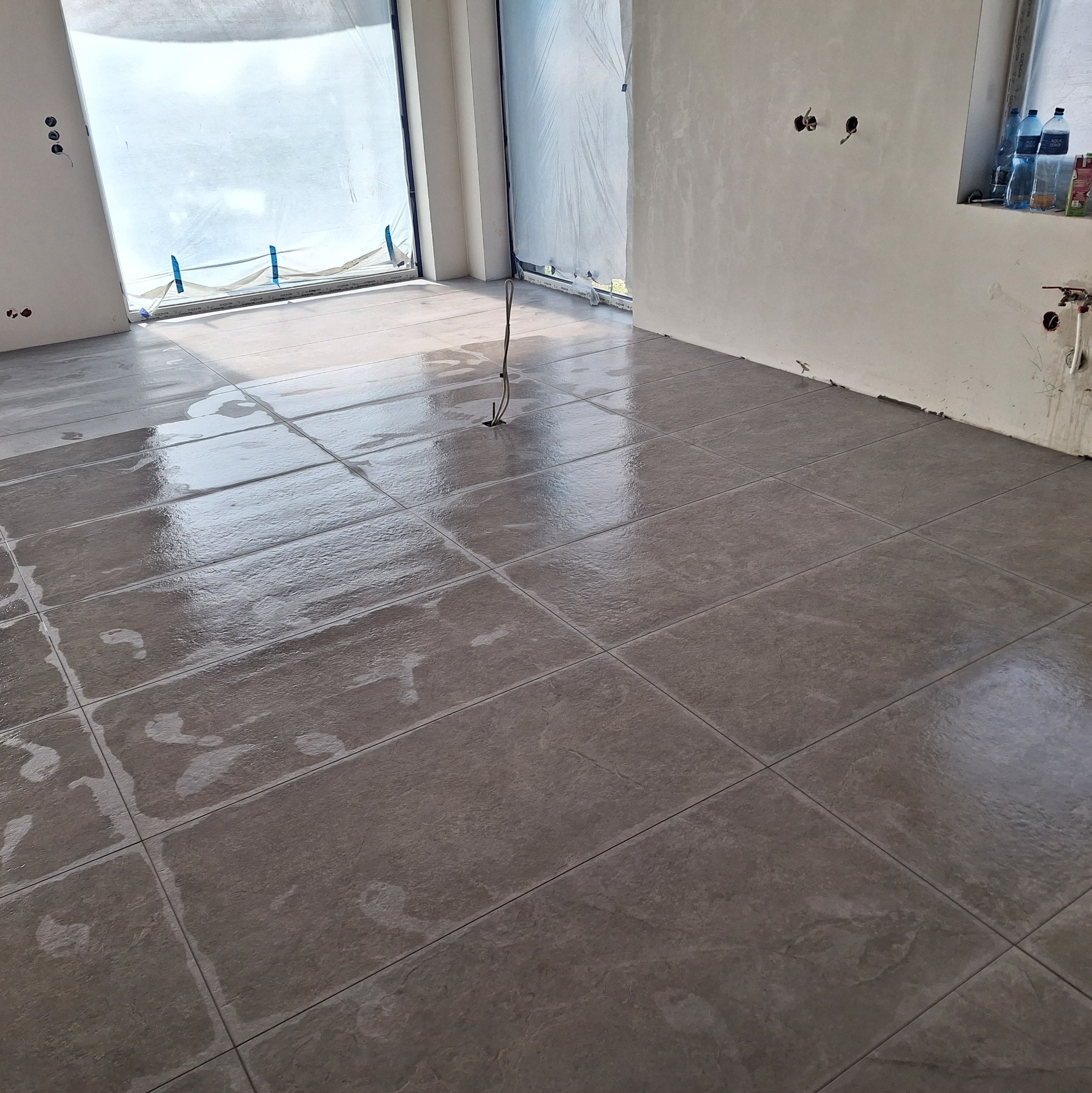 Świeżo położone, mokre, szare płytki podłogowe w pomieszczeniu w trakcie remontu, kable wystające z podłogi, okna zabezpieczone folią malarską.
