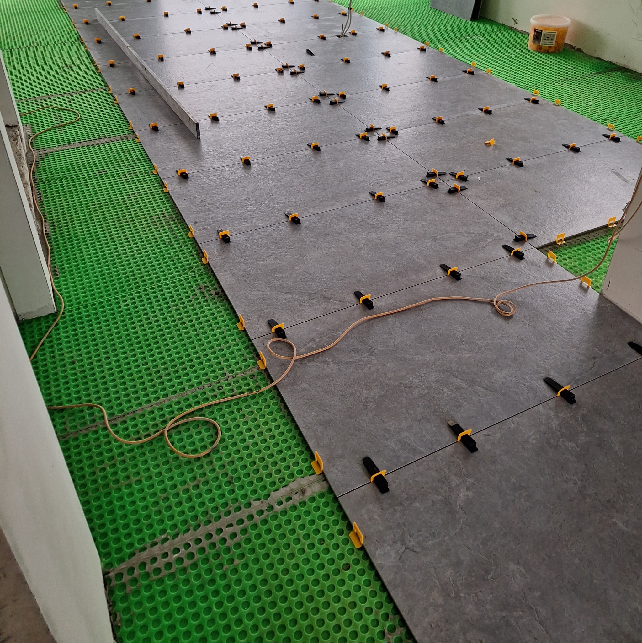 Szare płytki podłogowe z systemem poziomowania, ułożone na zielonej, perforowanej macie podkładowej, widoczny poziomica i pojemnik z klinami.