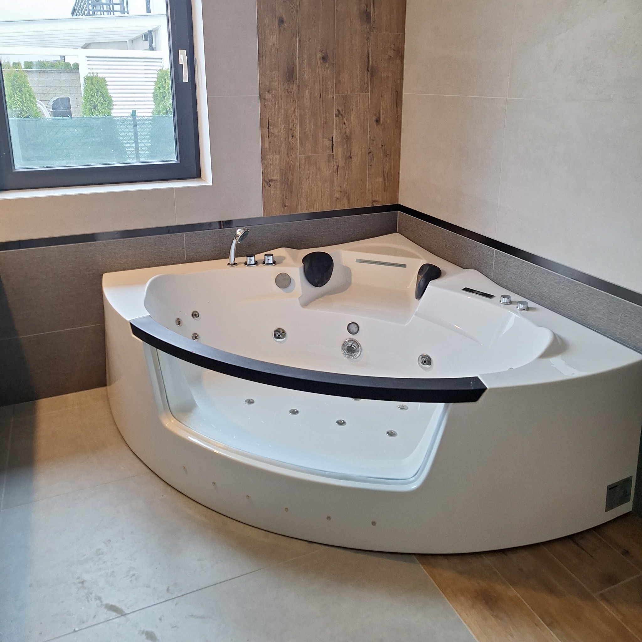 Nowoczesna, narożna wanna z hydromasażem w jasnej łazience, z widocznymi płytkami imitującymi drewno na ścianie i oknem z widokiem na zieleń.