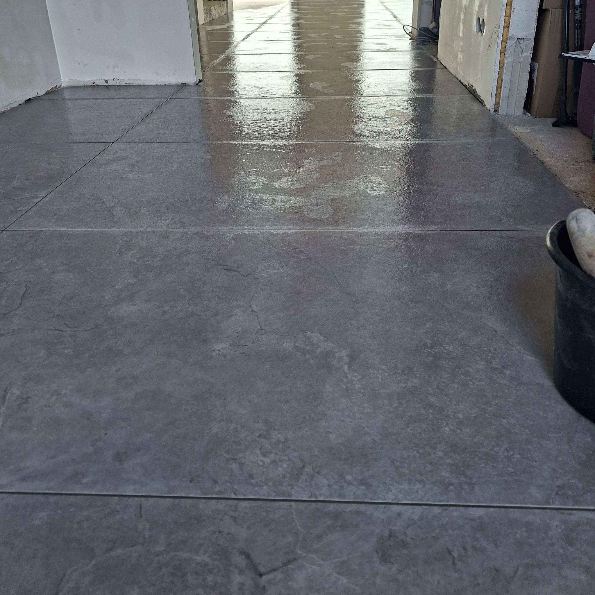 Świeżo ułożona, błyszcząca, szara podłoga z płytek imitujących kamień, z widocznymi śladami wilgoci i wiadrem z narzędziami w prawym dolnym rogu, w trakcie prac wykończeniowych.