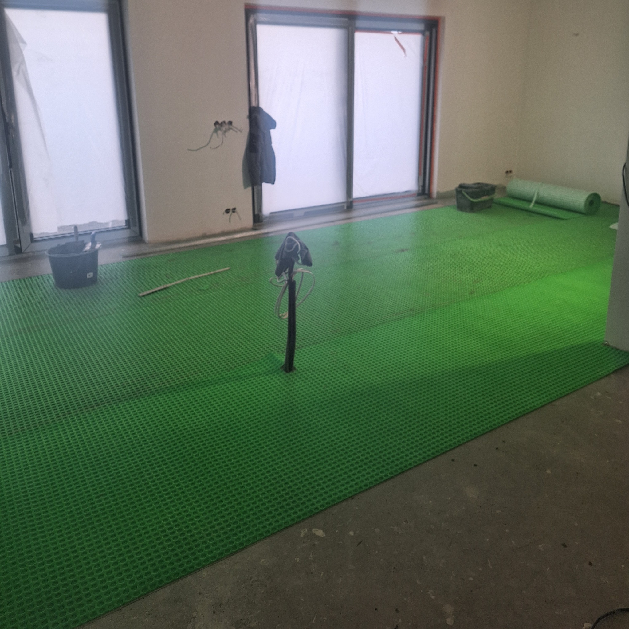 Przygotowanie podłoża pod wylewkę: zielona mata drenażowa ułożona na podłodze w surowym stanie, widoczne kable elektryczne wystające ze ściany, okna zabezpieczone folią.