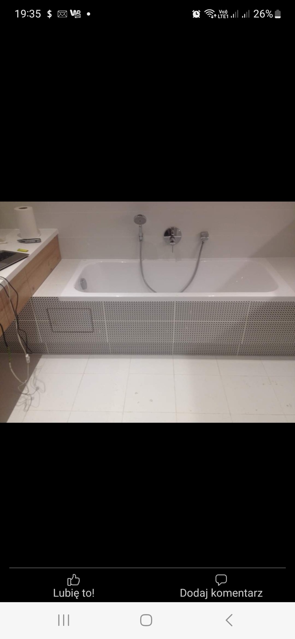 Biała wanna obudowana płytkami z drobnymi otworami, chromowana bateria i słuchawka prysznicowa na białej ścianie, obok blat z drewnopodobnym wykończeniem i laptopem.