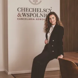 Portret kobiety w czarnym garniturze siedzącej na rogu biurka w biurze kancelarii adwokackiej Chechelscy & Wspólnicy, z logo firmy widocznym na ścianie.