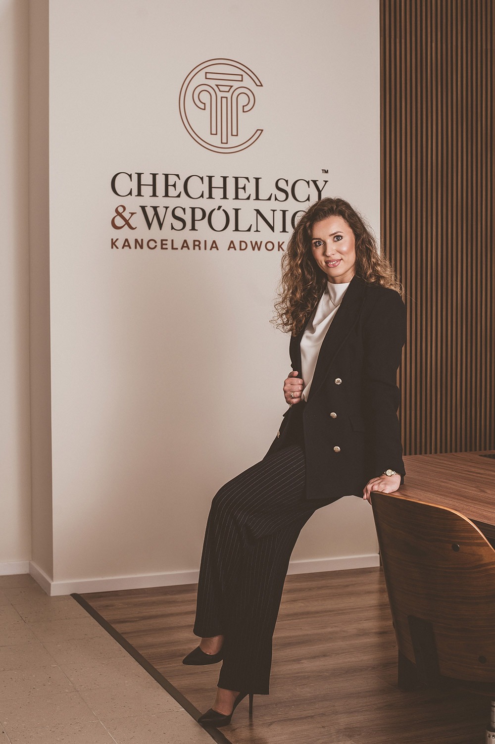 Portret kobiety w czarnym garniturze siedzącej na rogu biurka w biurze kancelarii adwokackiej Chechelscy & Wspólnicy, z logo firmy widocznym na ścianie.