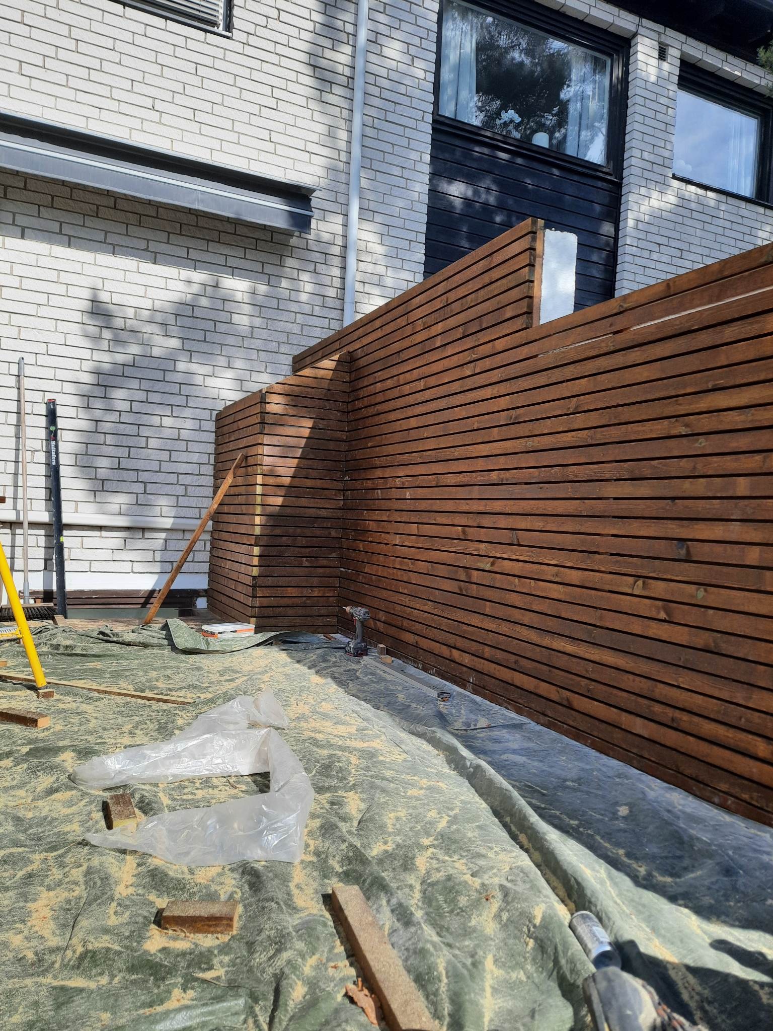 Montaż drewnianego ogrodzenia panelowego na zewnątrz budynku z białej cegły, widoczne narzędzia i poziomica, całość przykryta folią ochronną.