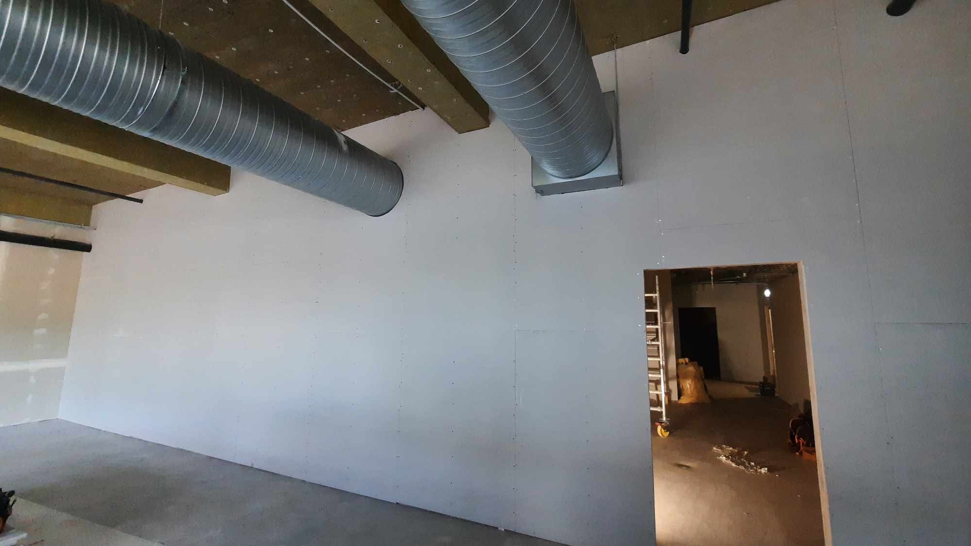 Wnętrze w trakcie prac wykończeniowych: ściany z płyt kartonowo-gipsowych, widoczne elementy wentylacji pod sufitem, otwór drzwiowy z drabiną w tle.