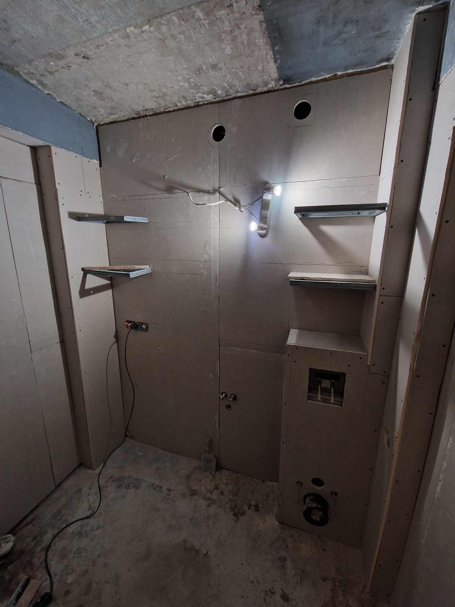Surowa łazienka w trakcie prac wykończeniowych, ściany wyłożone płytami gipsowo-kartonowymi, widoczne otwory na instalacje, metalowe wsporniki półek i wystająca instalacja elektryczna, sufit...