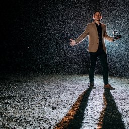 Movie ON - Mężczyzna w beżowej marynarce i ciemnych spodniach, stojący w silnym deszczu w nocy, trzyma aparat fotograficzny z lampą błyskową, oświetlony od dołu.