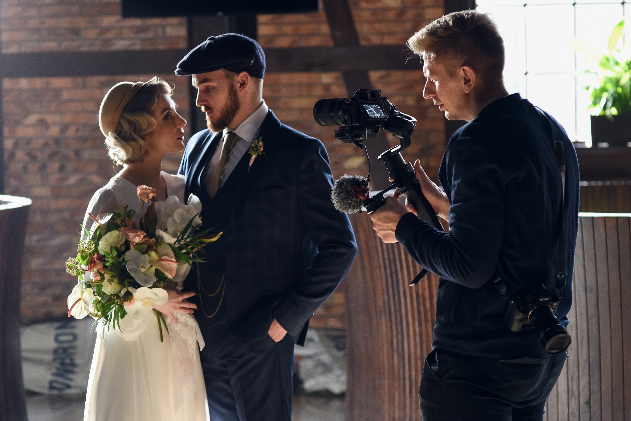 Kamerzysta filmuje parę młodą w stylu retro, używając kamery na stabilizatorze z mikrofonem zewnętrznym, w tle ceglana ściana i drewniane elementy.