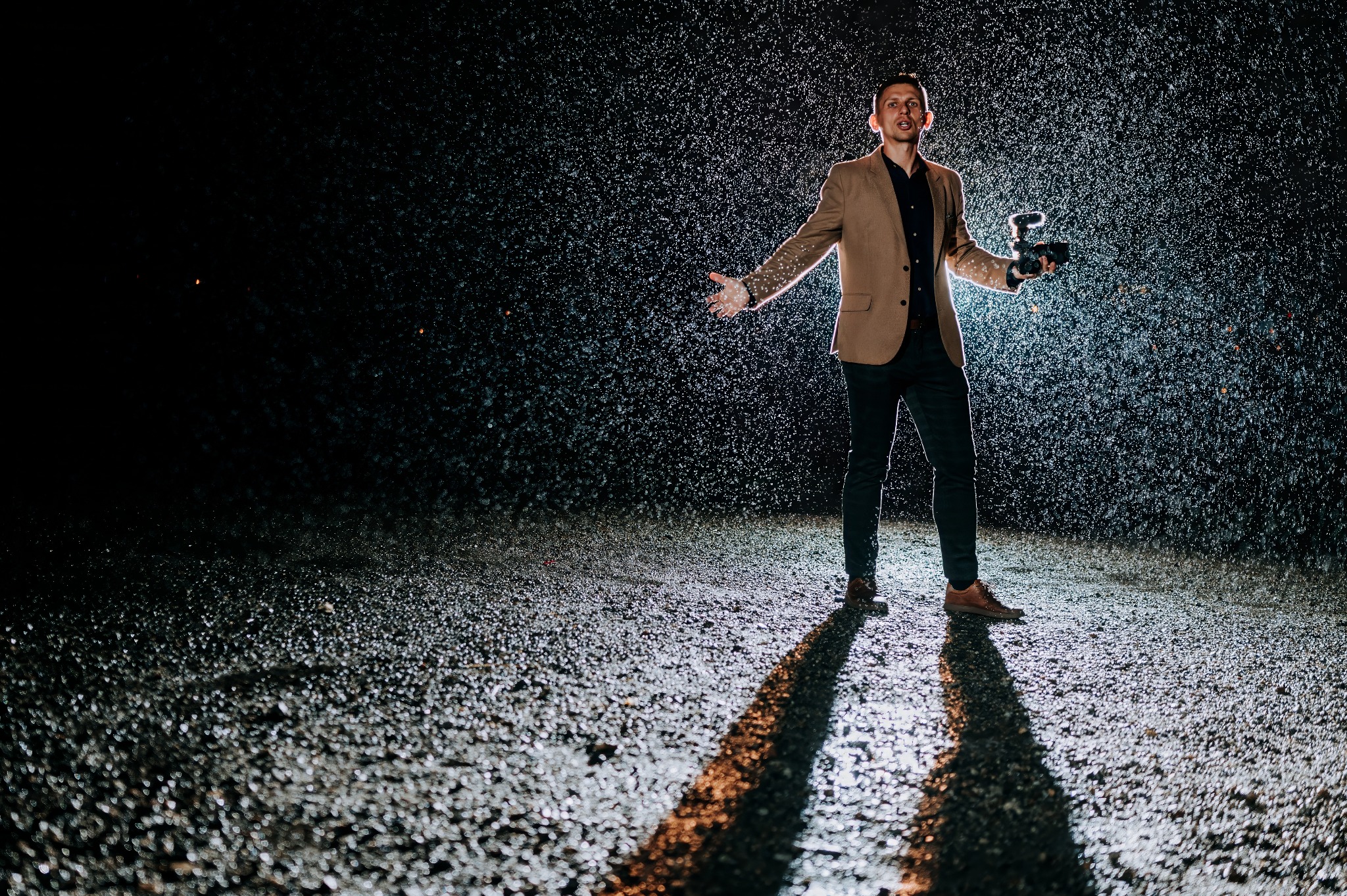 Mężczyzna w beżowej marynarce i ciemnych spodniach, stojący w silnym deszczu w nocy, trzyma aparat fotograficzny z lampą błyskową, oświetlony od dołu.