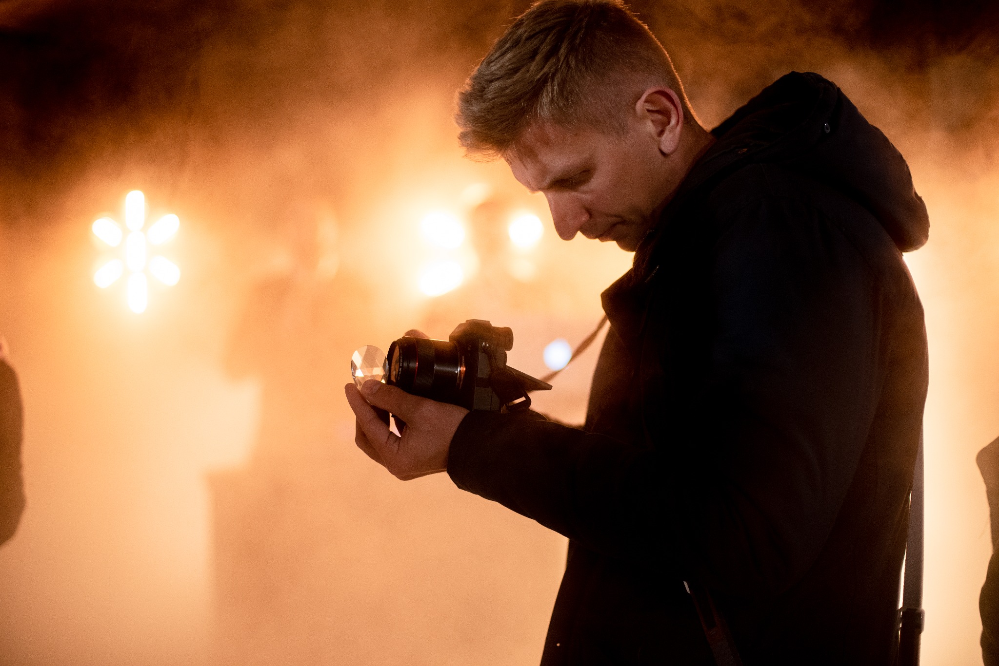 Młody mężczyzna w ciemnej kurtce, pochylony, trzyma w dłoniach aparat z przymocowanym pryzmatem, sprawdzając ustawienia, na tle rozproszonego pomarańczowego światła i sylwetek innych osób.