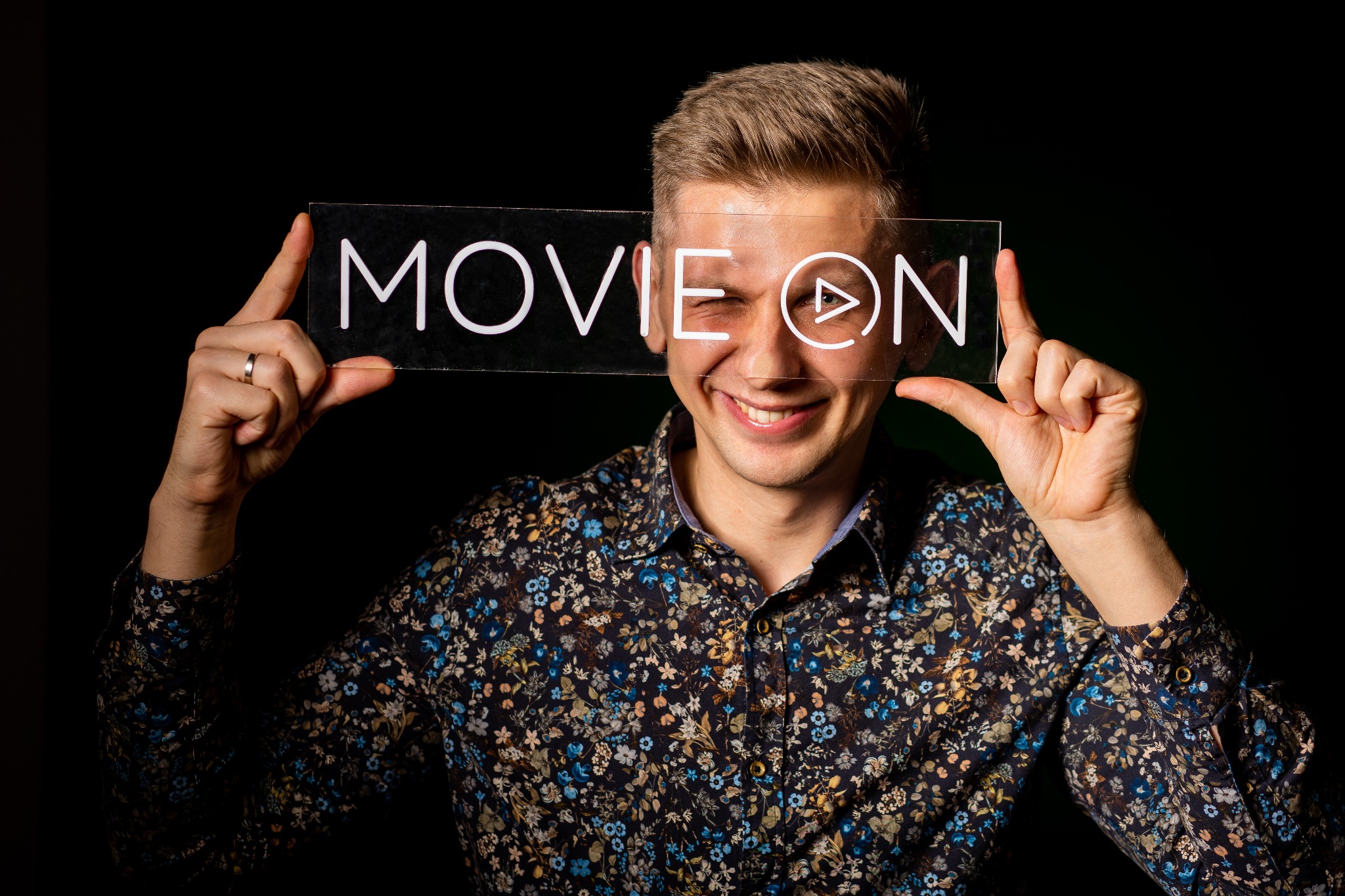 Uśmiechnięty mężczyzna z Krakowa mruga okiem, trzymając przed twarzą przezroczystą tabliczkę z napisem 'MOVIE ON' i symbolem odtwarzania w kółku na czarnym tle.