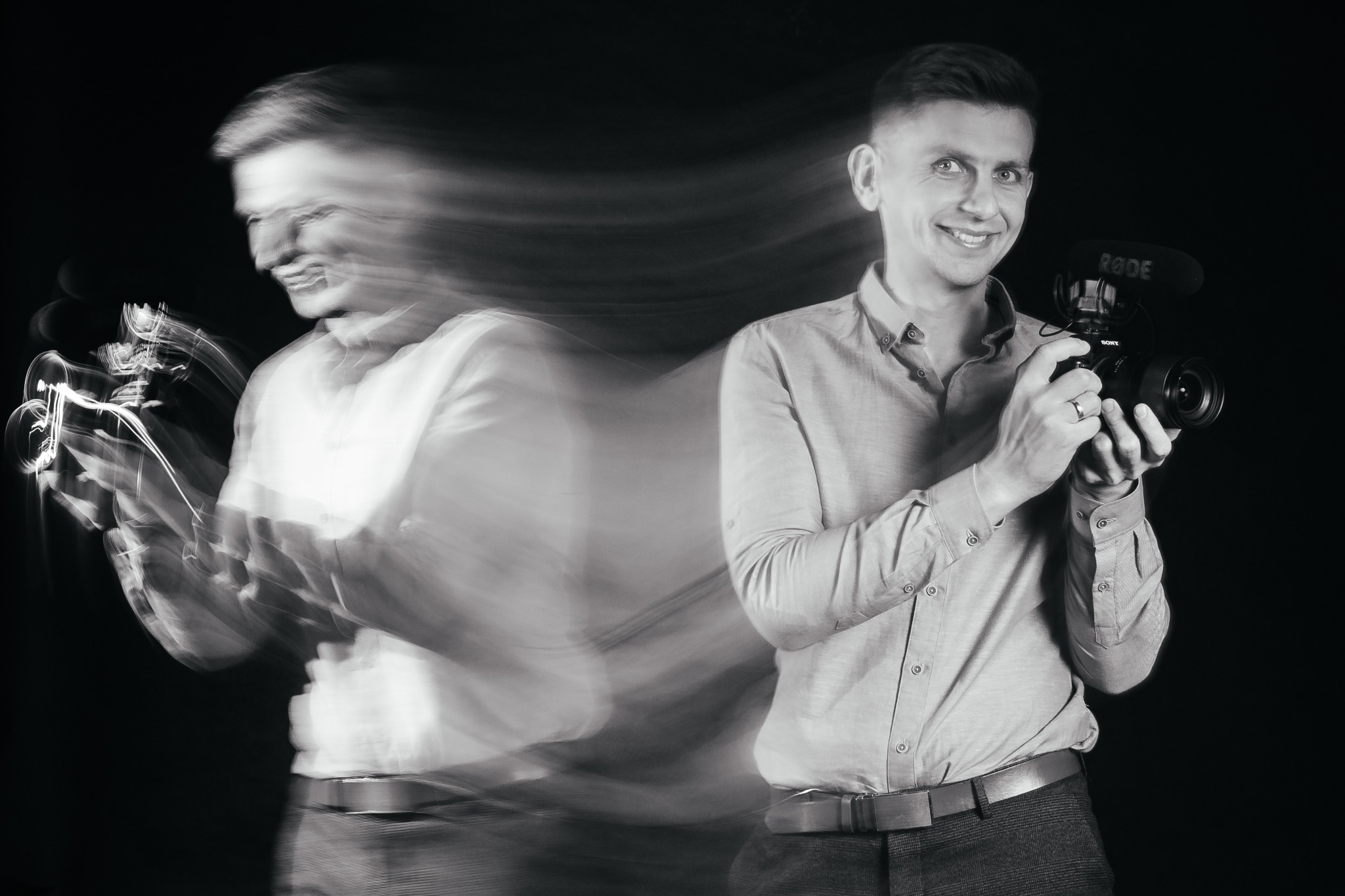 Czarno-białe zdjęcie uśmiechniętego mężczyzny trzymającego kamerę Sony z mikrofonem Rode, z efektem rozmycia ruchu po lewej stronie.