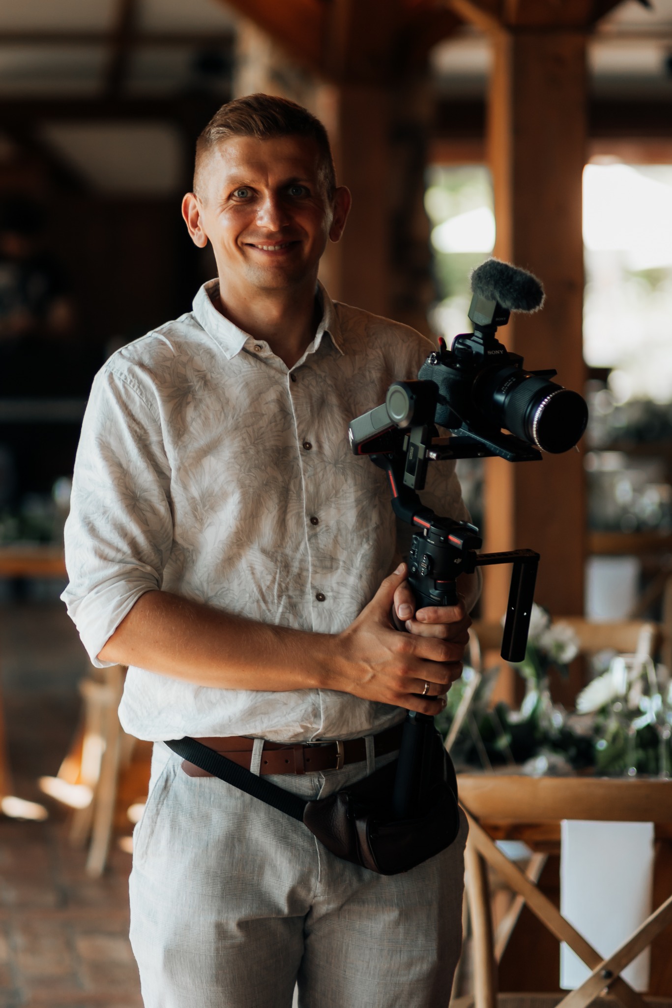 Uśmiechnięty mężczyzna w koszuli w palmy trzyma profesjonalny aparat z mikrofonem na stabilizatorze, w tle drewniana konstrukcja i dekoracje kwiatowe.