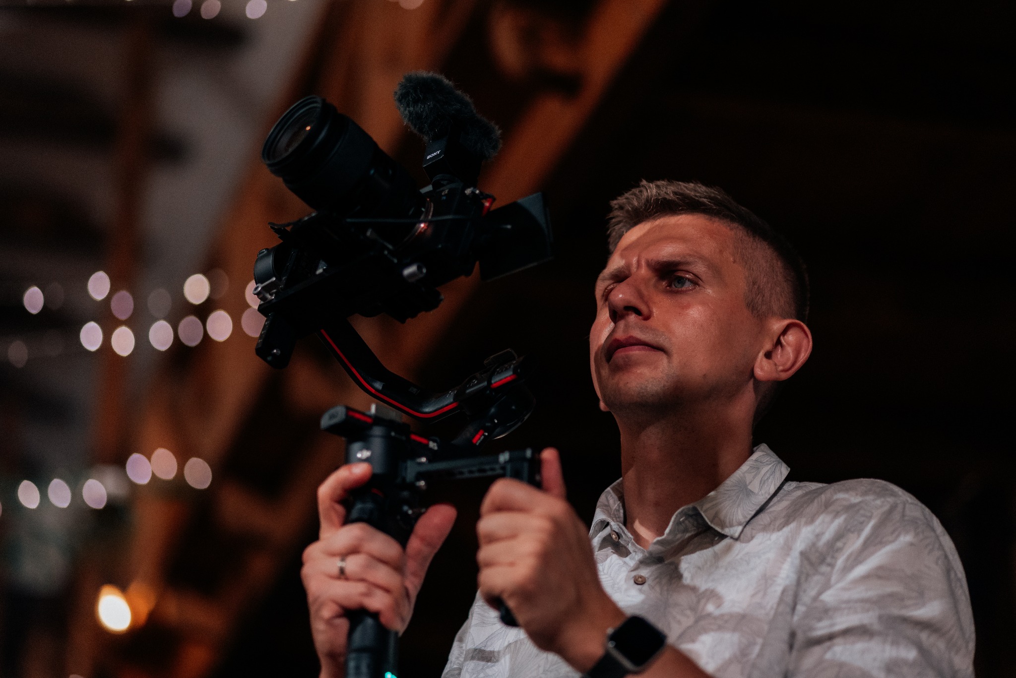 Profesjonalny kamerzysta z gimbalem i mikrofonem kierunkowym, skupiony wzrok, w tle rozmyte światła.