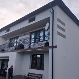 Pawel Skuza usługi remontowo budowlane - Dwukondygnacyjny dom z jasną elewacją, balkonem z ozdobną balustradą, satelitą i dekoracyjnymi pasami. Mężczyzna z psem na chodniku.