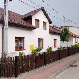 Pawel Skuza usługi remontowo budowlane - Biały dom jednorodzinny z brązowym dachem i drewnianymi oknami, widok z ulicy z brązową drewnianą barierką, z żółtą skrzynką gazową na elewacji.