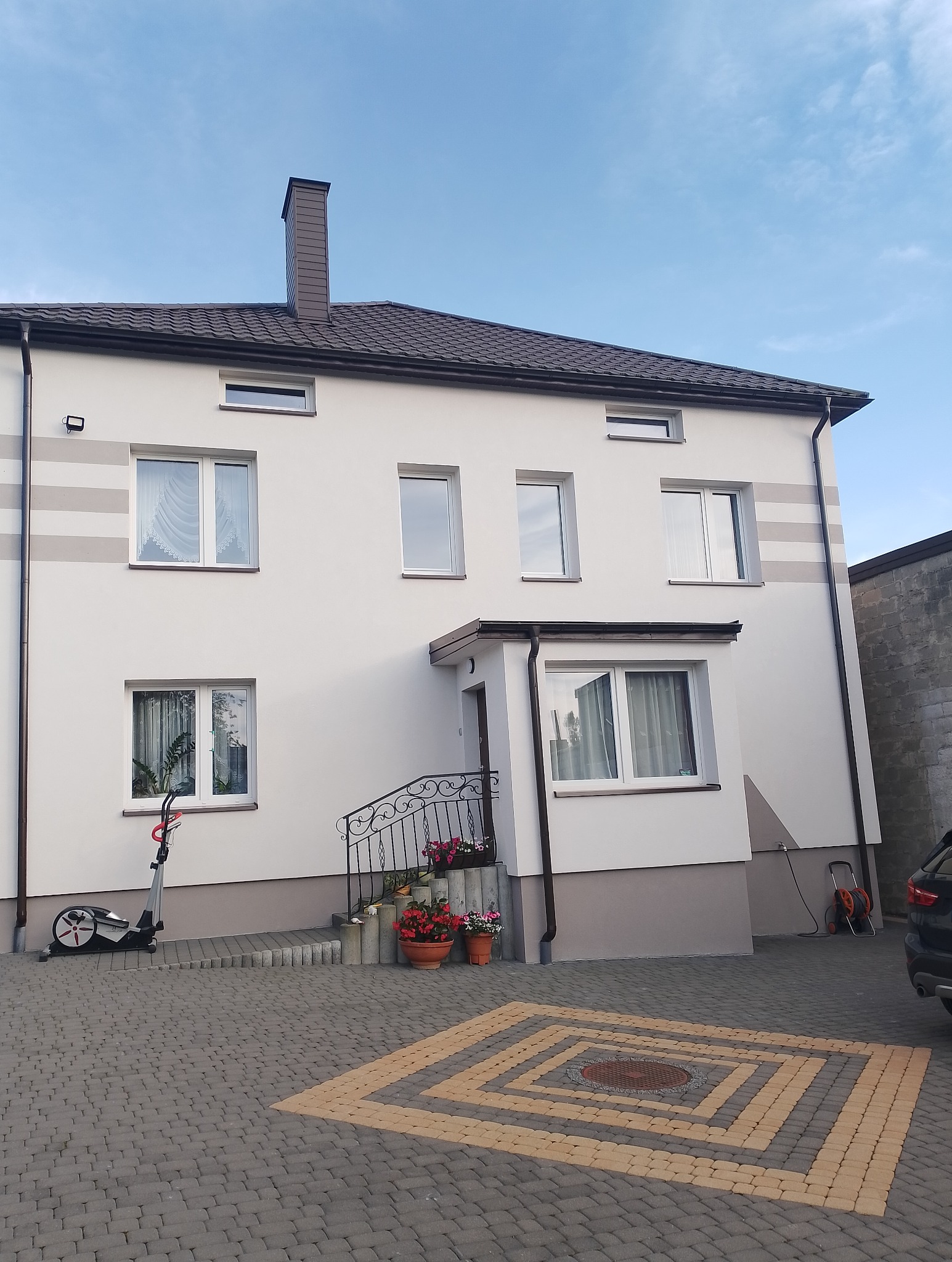 Dwupiętrowy dom z jasną elewacją, brązowym dachem i rynnami, widoczny z poziomu kostki brukowej z dekoracyjnym wzorem, obok stoi rower treningowy.