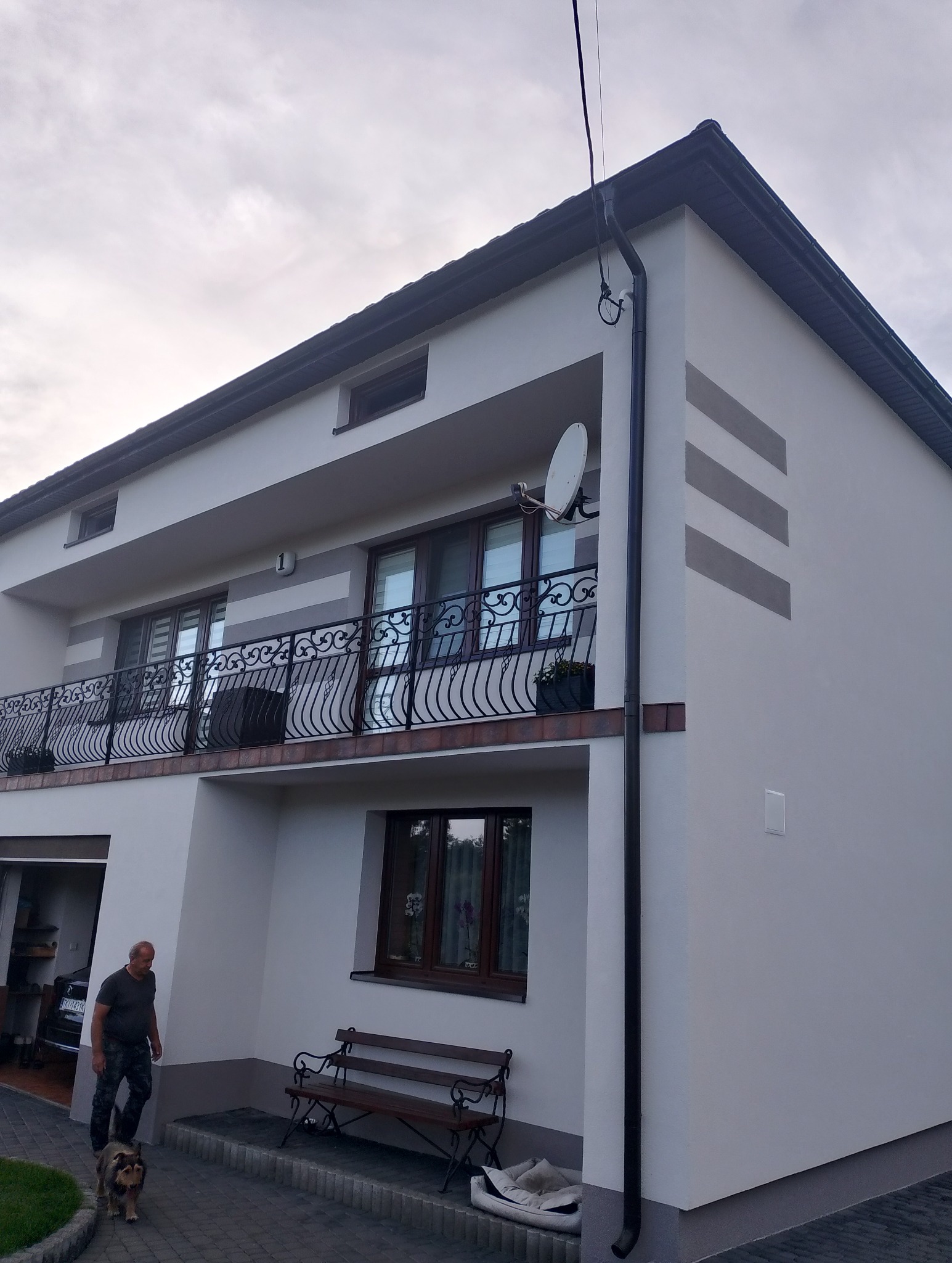 Dwukondygnacyjny dom z jasną elewacją, balkonem z ozdobną balustradą, satelitą i dekoracyjnymi pasami. Mężczyzna z psem na chodniku.