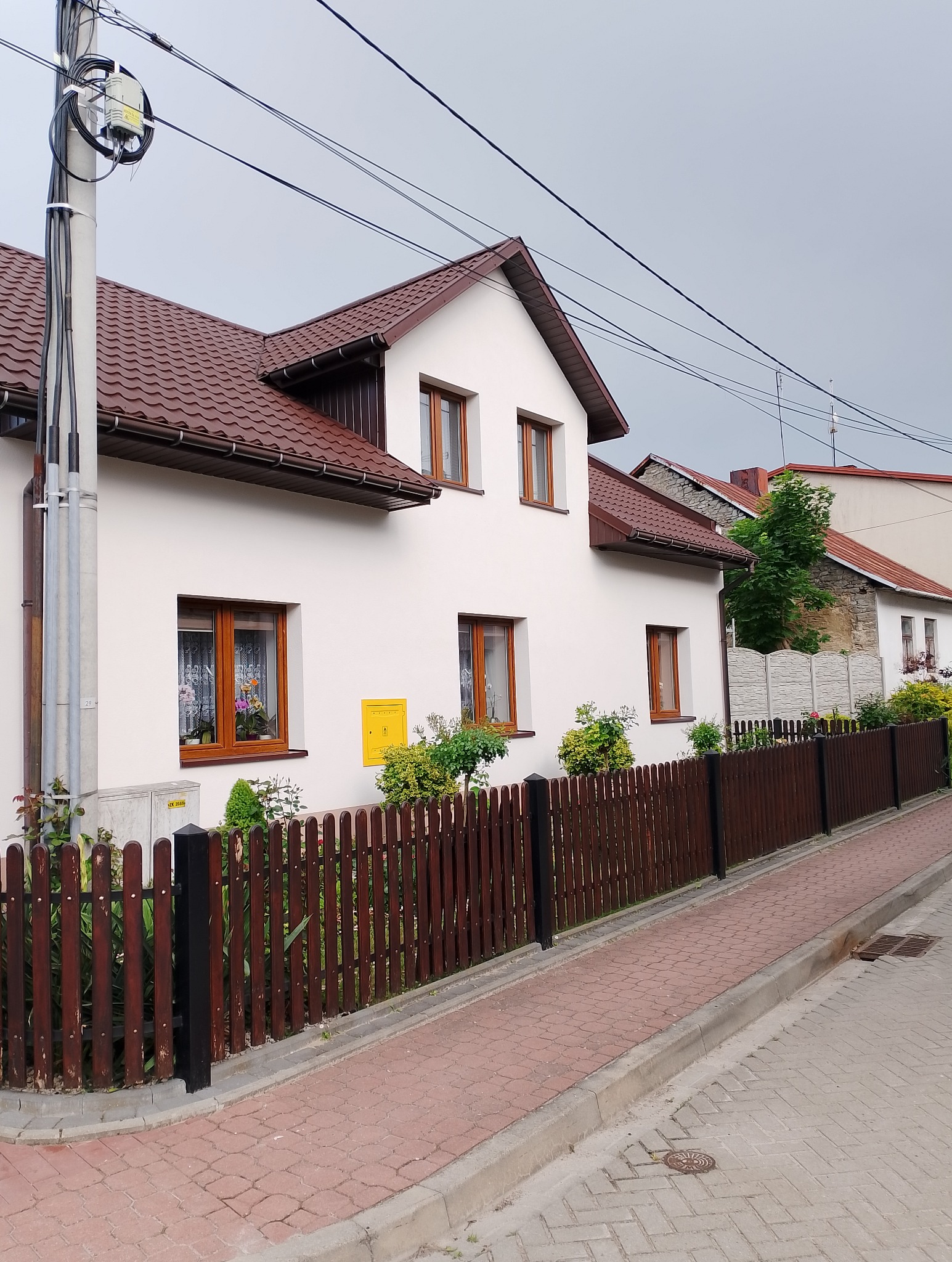 Biały dom jednorodzinny z brązowym dachem i drewnianymi oknami, widok z ulicy z brązową drewnianą barierką, z żółtą skrzynką gazową na elewacji.