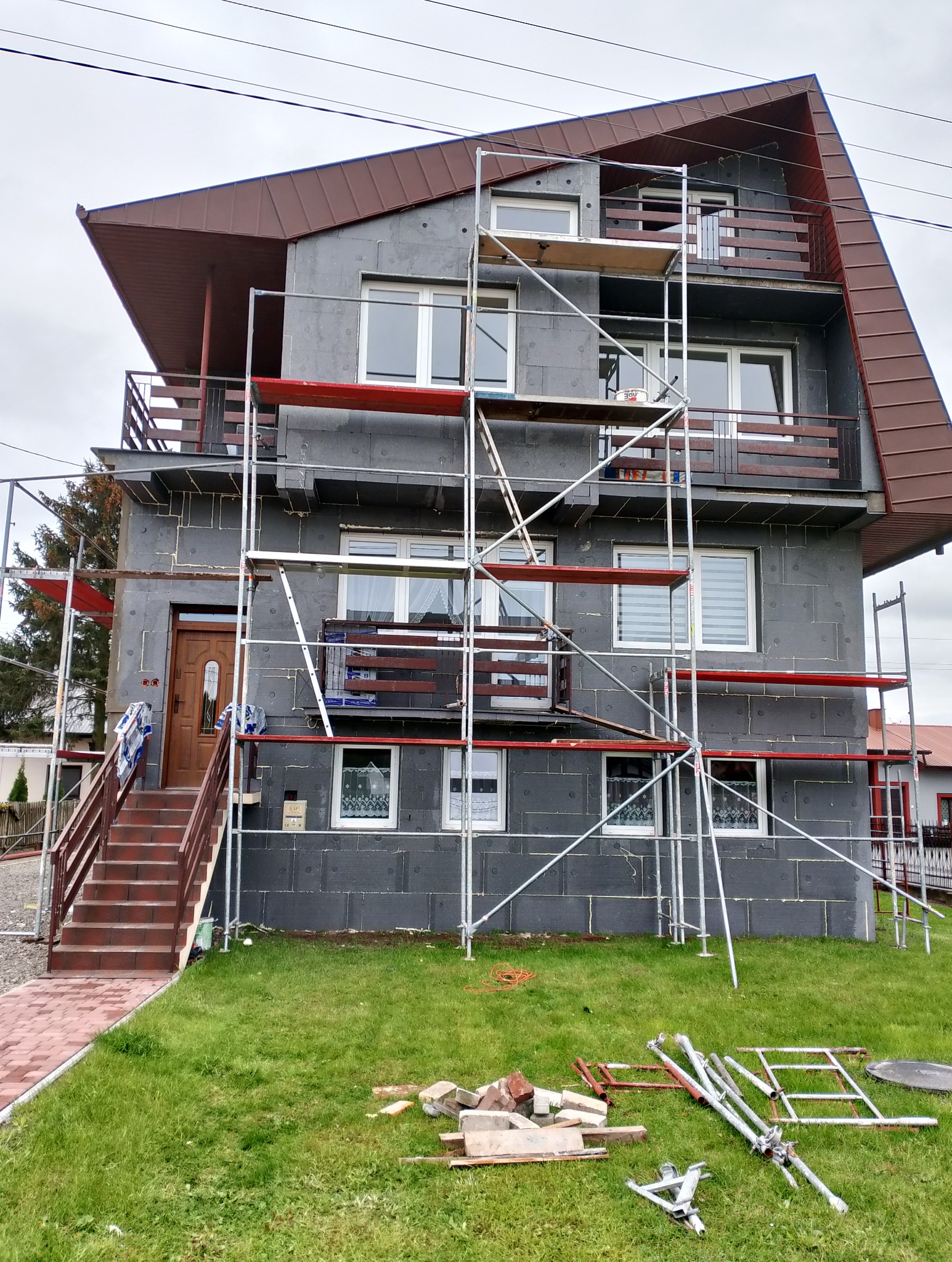 Dom jednorodzinny w trakcie ocieplania, widoczne rusztowania, styropian na ścianach, brązowy dach i schody wejściowe z czerwonej cegły