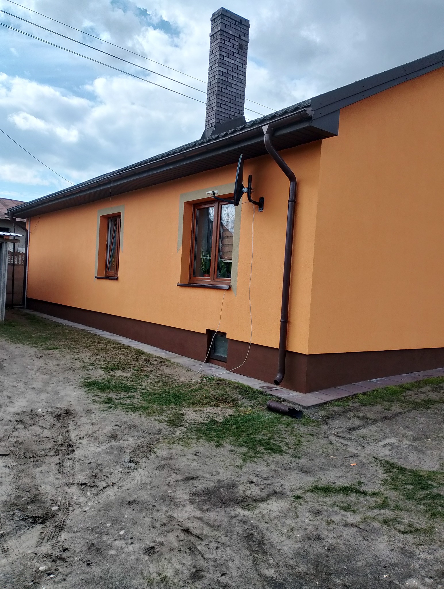 Ocieplony dom z pomarańczową elewacją, brązowym cokołem i ceglanym kominem, widoczna antena satelitarna na ścianie.