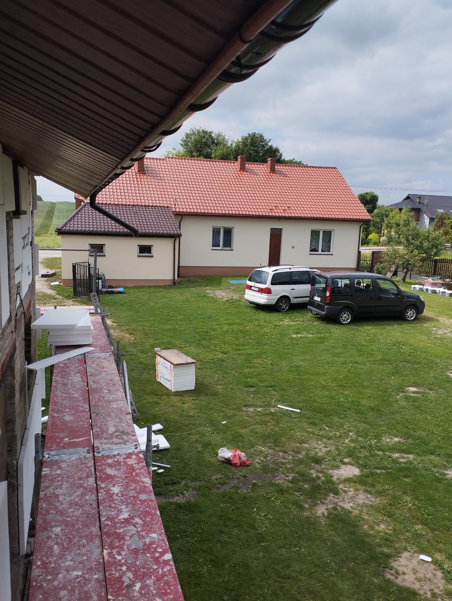 Dom jednorodzinny z częściowo ocieploną ścianą, widok z rusztowania, styropianowe płyty do ocieplenia oraz dwa samochody na trawniku w tle.