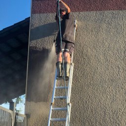 Hugo clean - Mężczyzna na drabinie w gumowych butach czyści elewację domu za pomocą myjki ciśnieniowej, widoczny strumień wody i różnica w kolorze czyszczonej powierzchni.