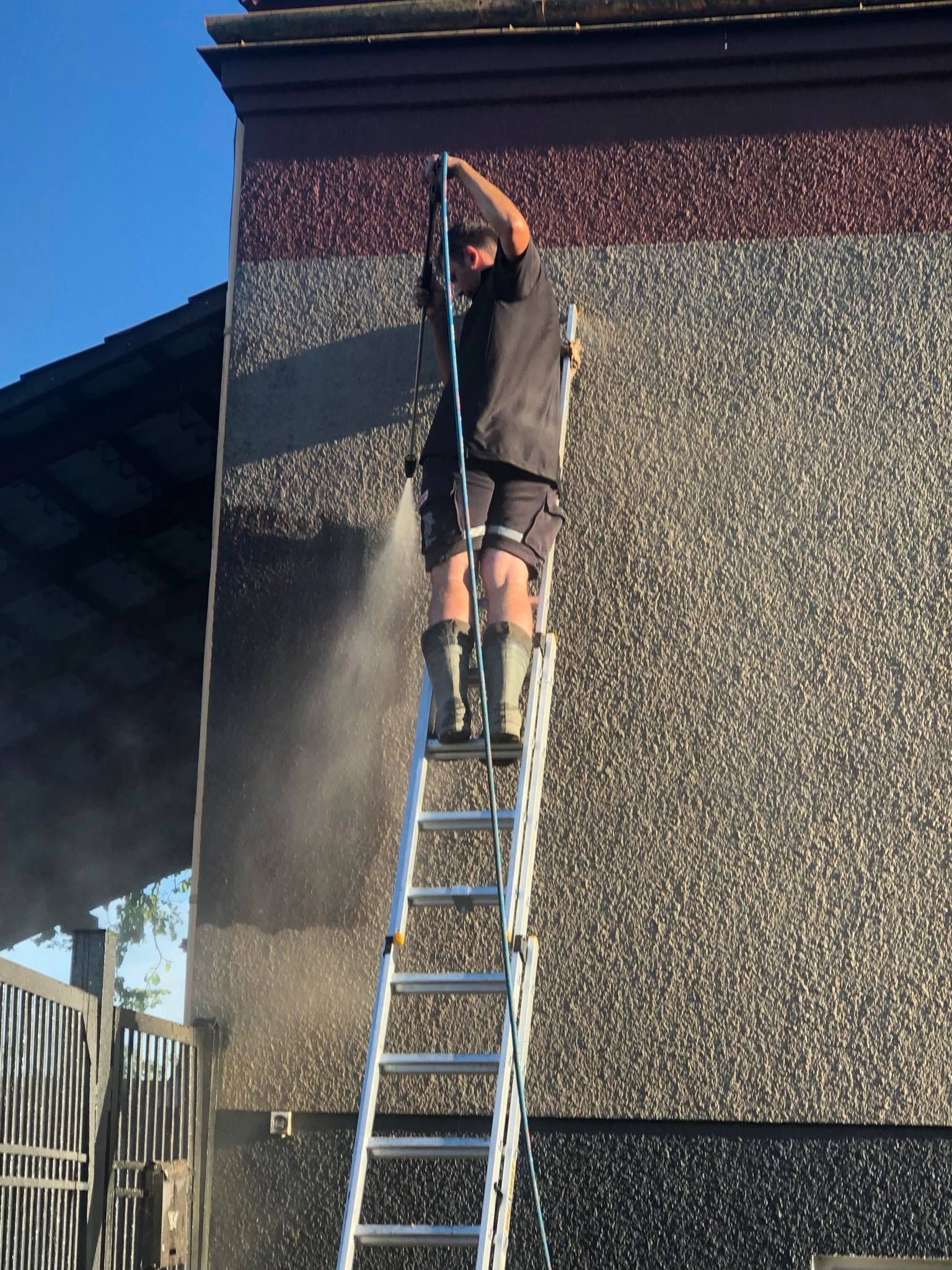 Mężczyzna na drabinie w gumowych butach czyści elewację domu za pomocą myjki ciśnieniowej, widoczny strumień wody i różnica w kolorze czyszczonej powierzchni.