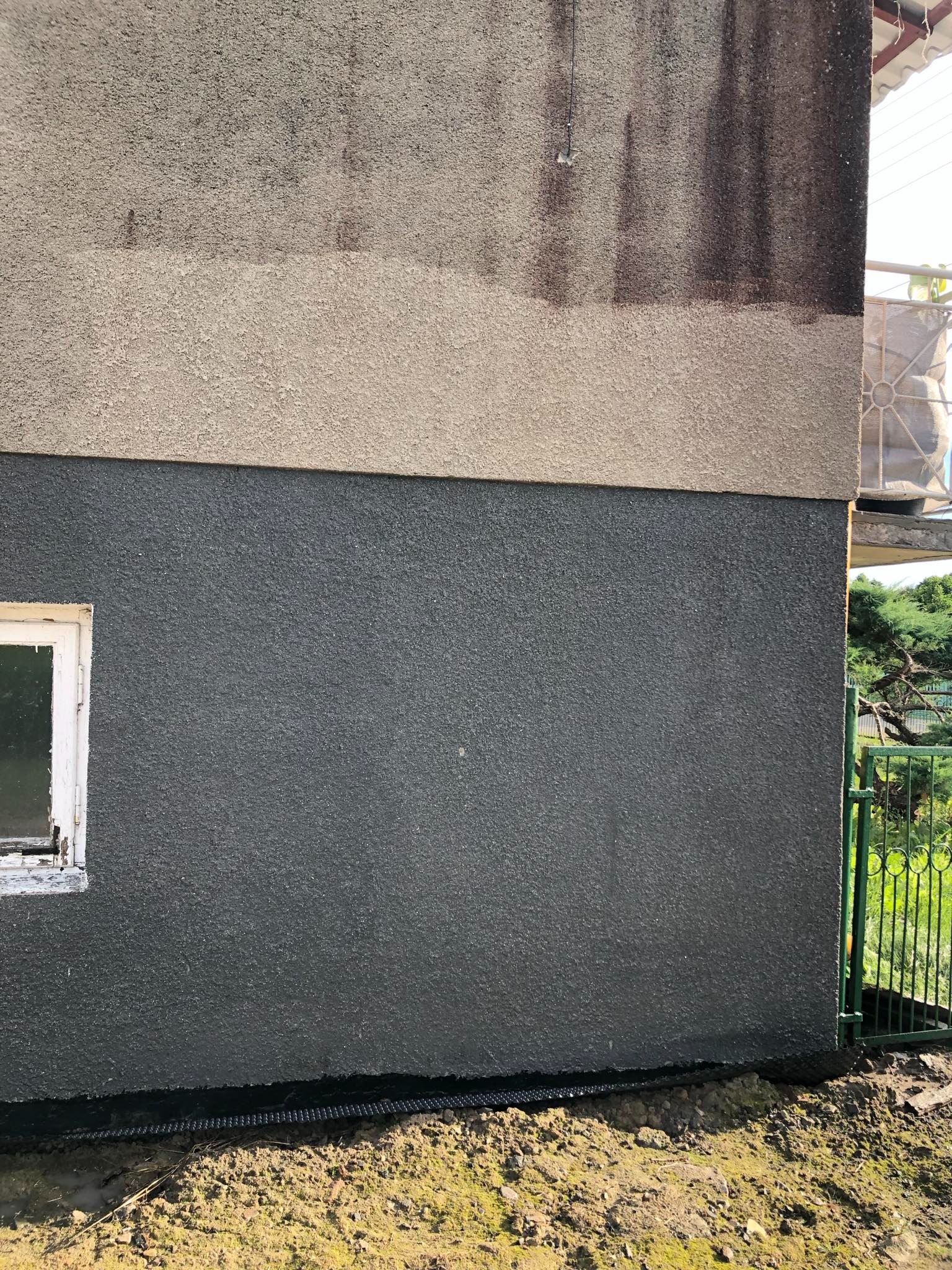 Fragment elewacji budynku z widocznym podziałem na dwie warstwy tynku: górna, jaśniejsza, z widocznymi zaciekami i zabrudzeniami, oraz dolna, ciemnoszara, świeżo nałożona. Przy elewacji widoczna...