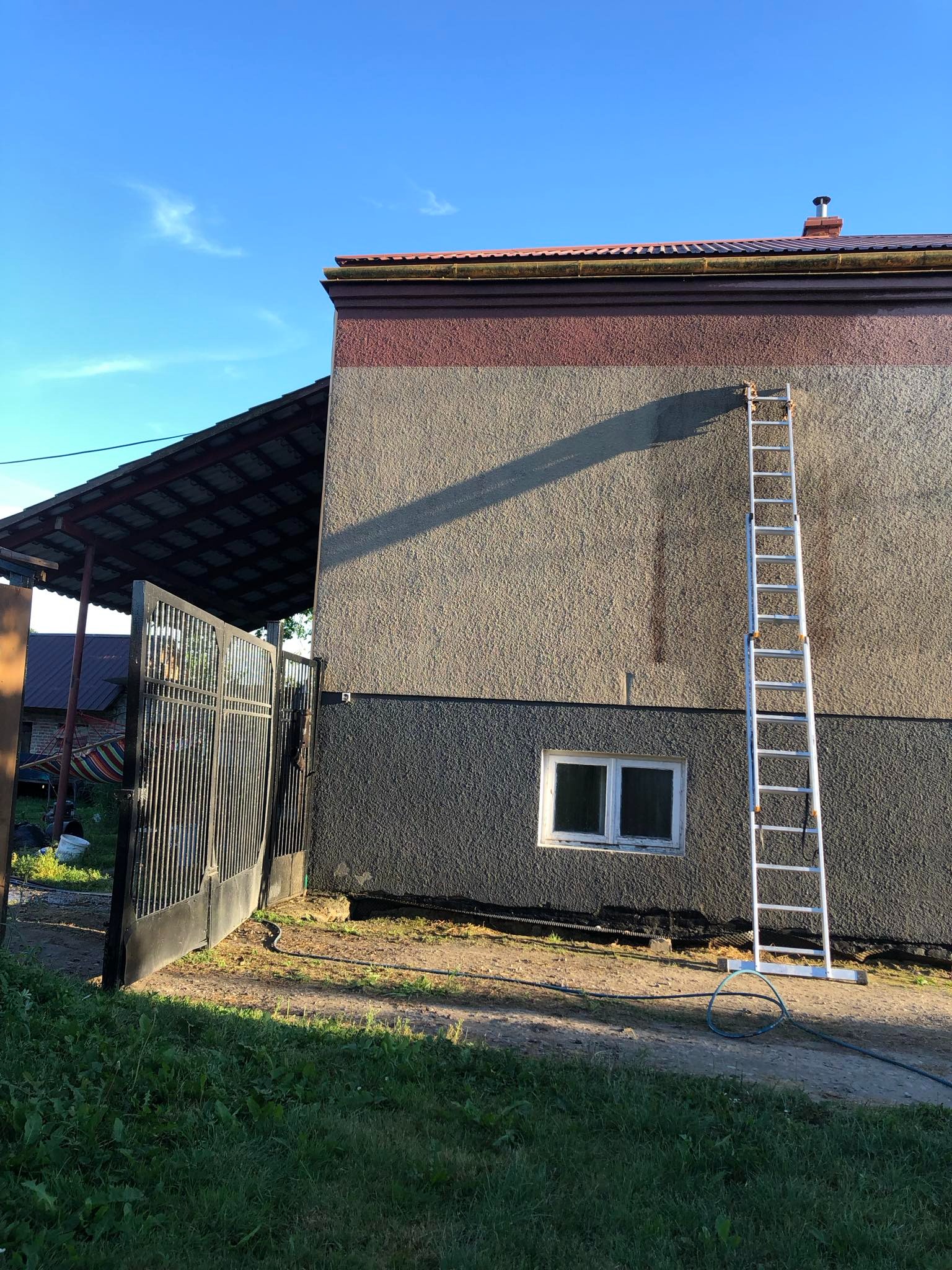 Dwukolorowa elewacja domu z teksturą, z metalową drabiną opartą o ścianę, widoczna brama wjazdowa i fragment trawnika.