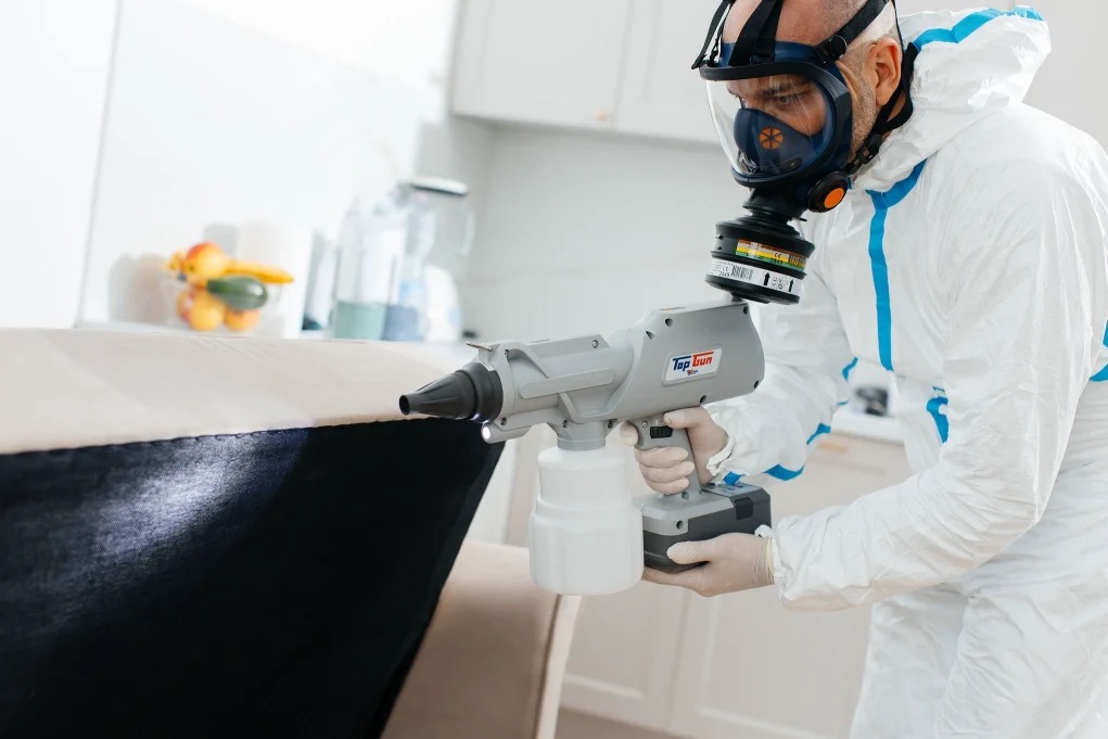 Pracownik w kombinezonie ochronnym i masce, używa zamgławiacza ULV Top Gun do dezynfekcji tapicerki meblowej w jasnym wnętrzu.
