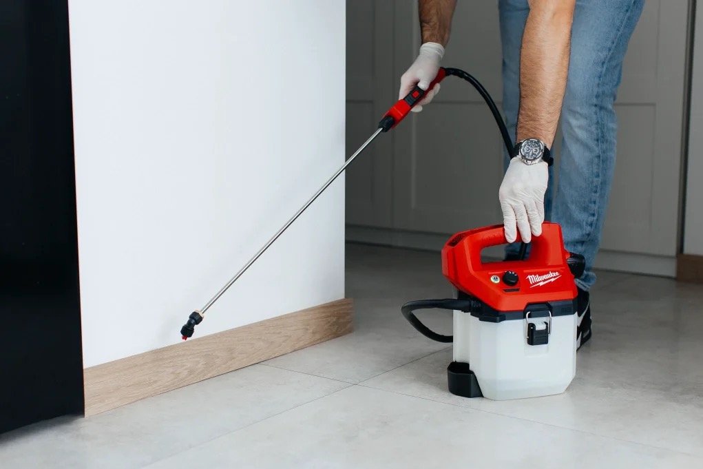 Oprysk ciśnieniowy z użyciem czerwonego urządzenia Milwaukee, aplikacja preparatu na ścianę przy podłodze, pracownik w rękawicach i jeansach, widoczny zegarek na ręce.