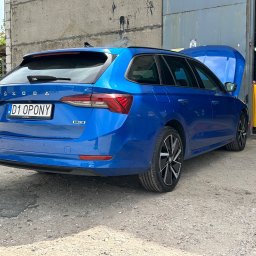MOTOINVEST Paweł Nowrotek - Niebieska Skoda Octavia kombi z otwartą maską stoi przed warsztatem; w tle widoczny żółty automat do obsługi klimatyzacji samochodowej Texa 707 Touch R-1234YF.